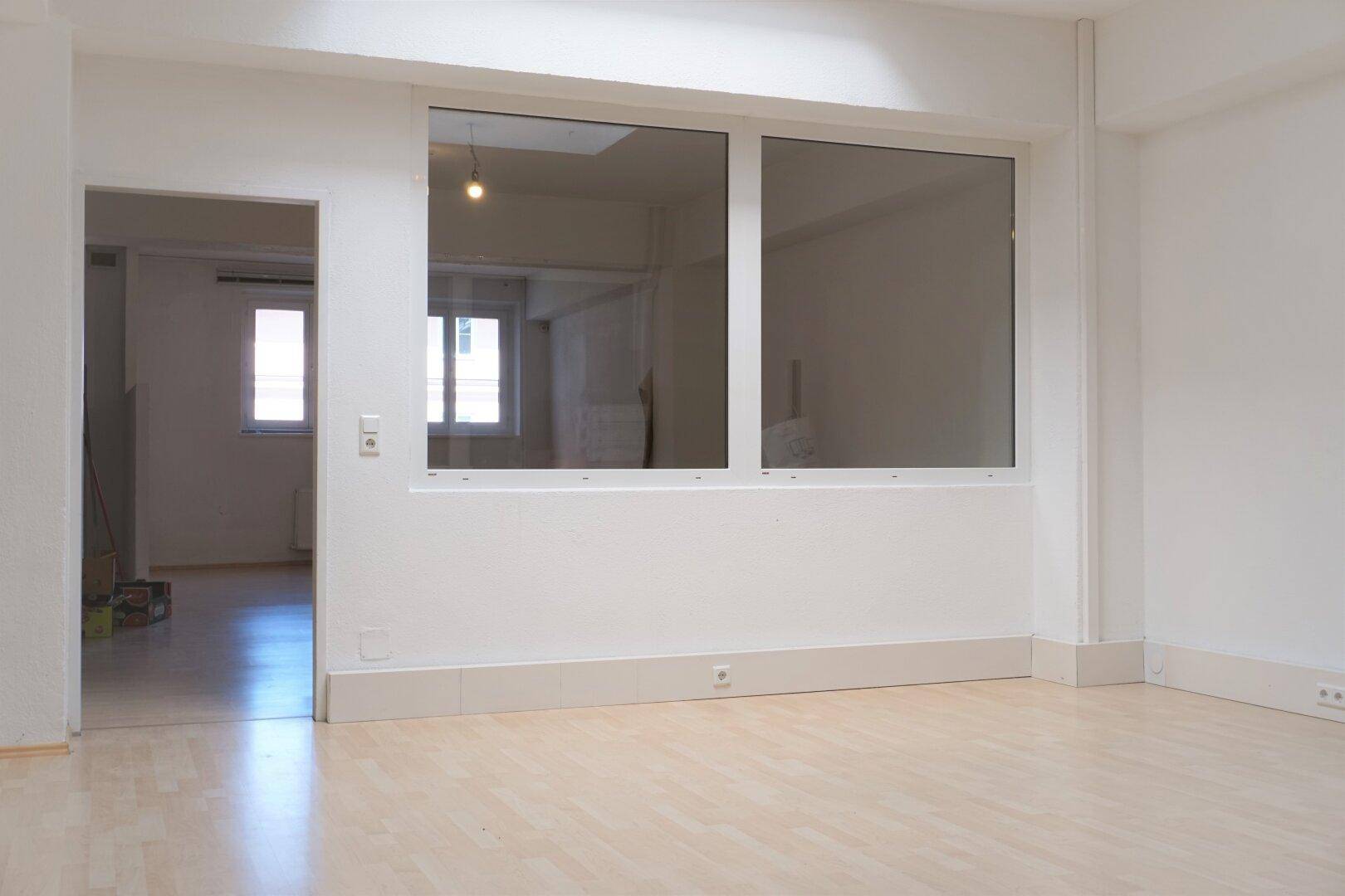Geräumiger Raum mit hellem Holzboden, weißen Wänden und großem Fenster, das eine Verbindung zum Nebenraum schafft.