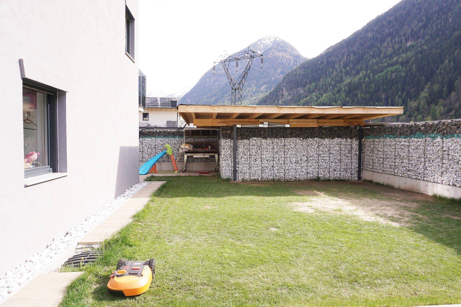 Gepflegter Gartenbereich mit Rasen, Grillplatz und schattenspendendem Unterstand, umgeben von Bergen.