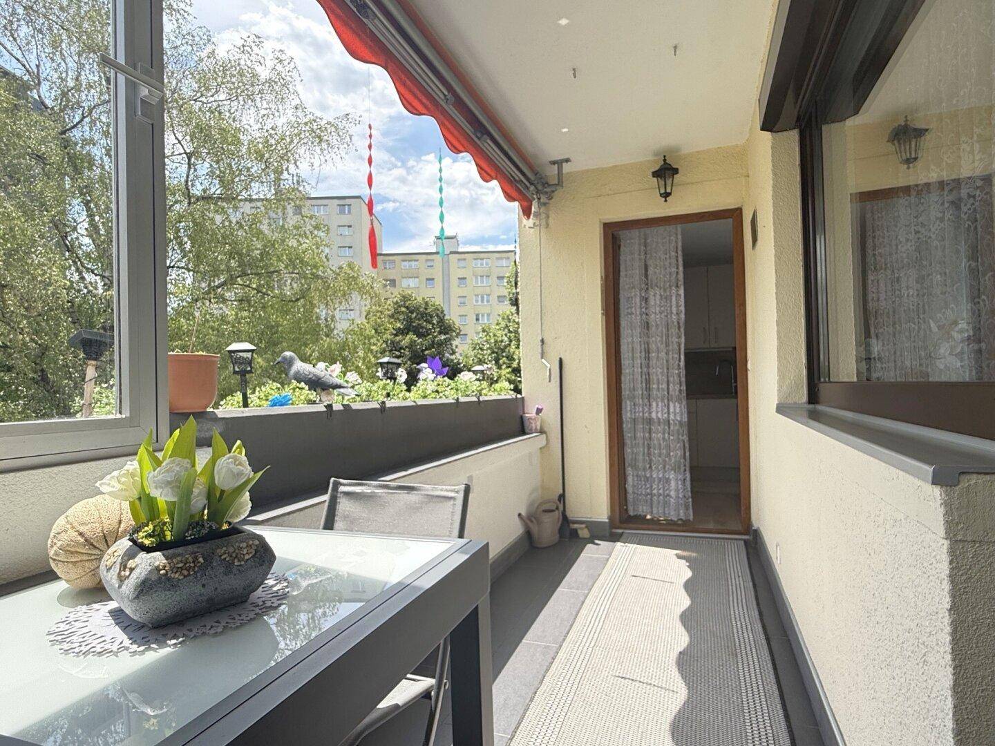Verglaster Balkon mit Sitzgelegenheiten und Blick auf die umliegenden Gebäude und Grünflächen.