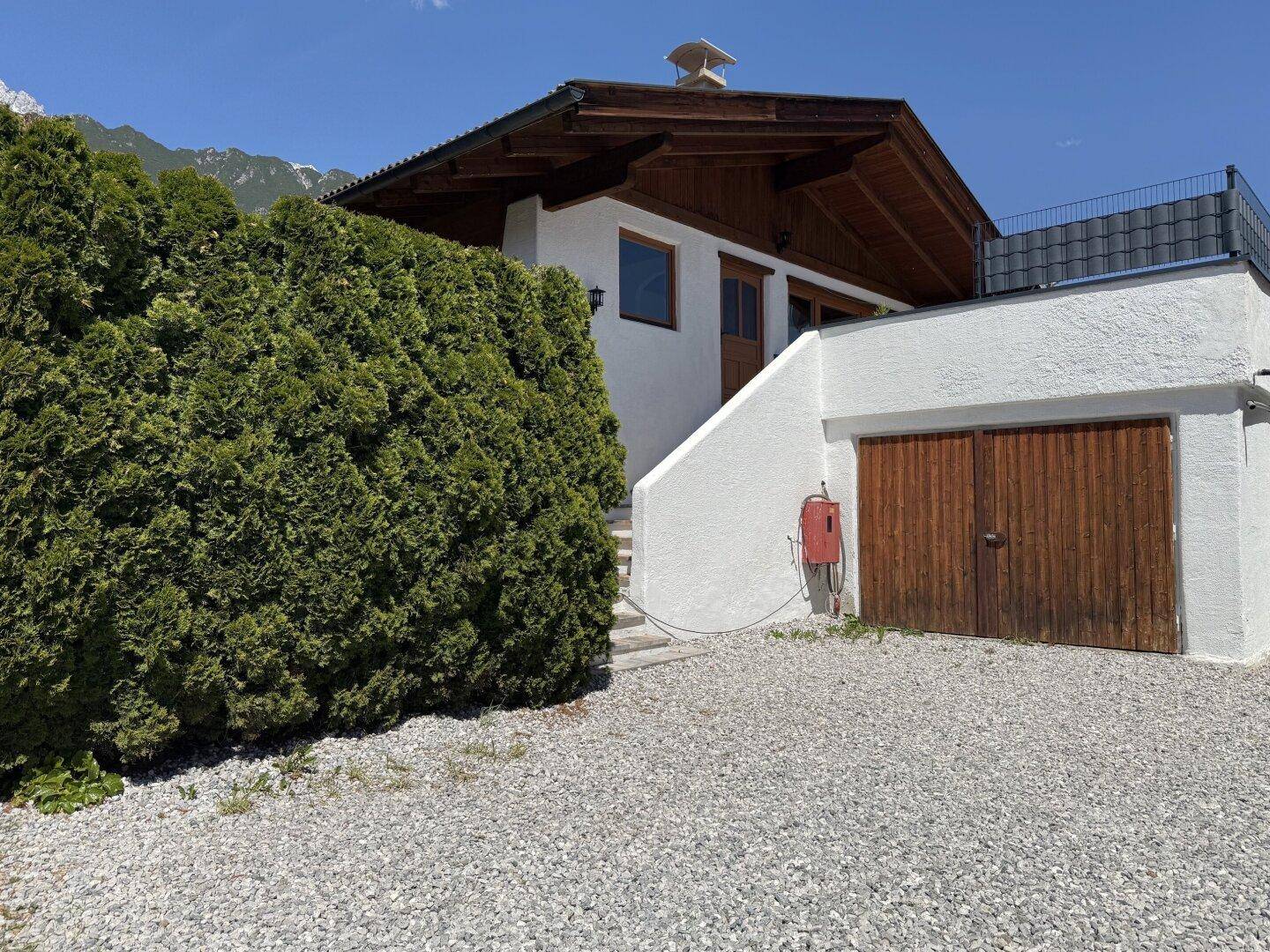 Außenansicht des Hauses mit Garage, Außentreppe und üppiger Hecke vor einer Bergkulisse.