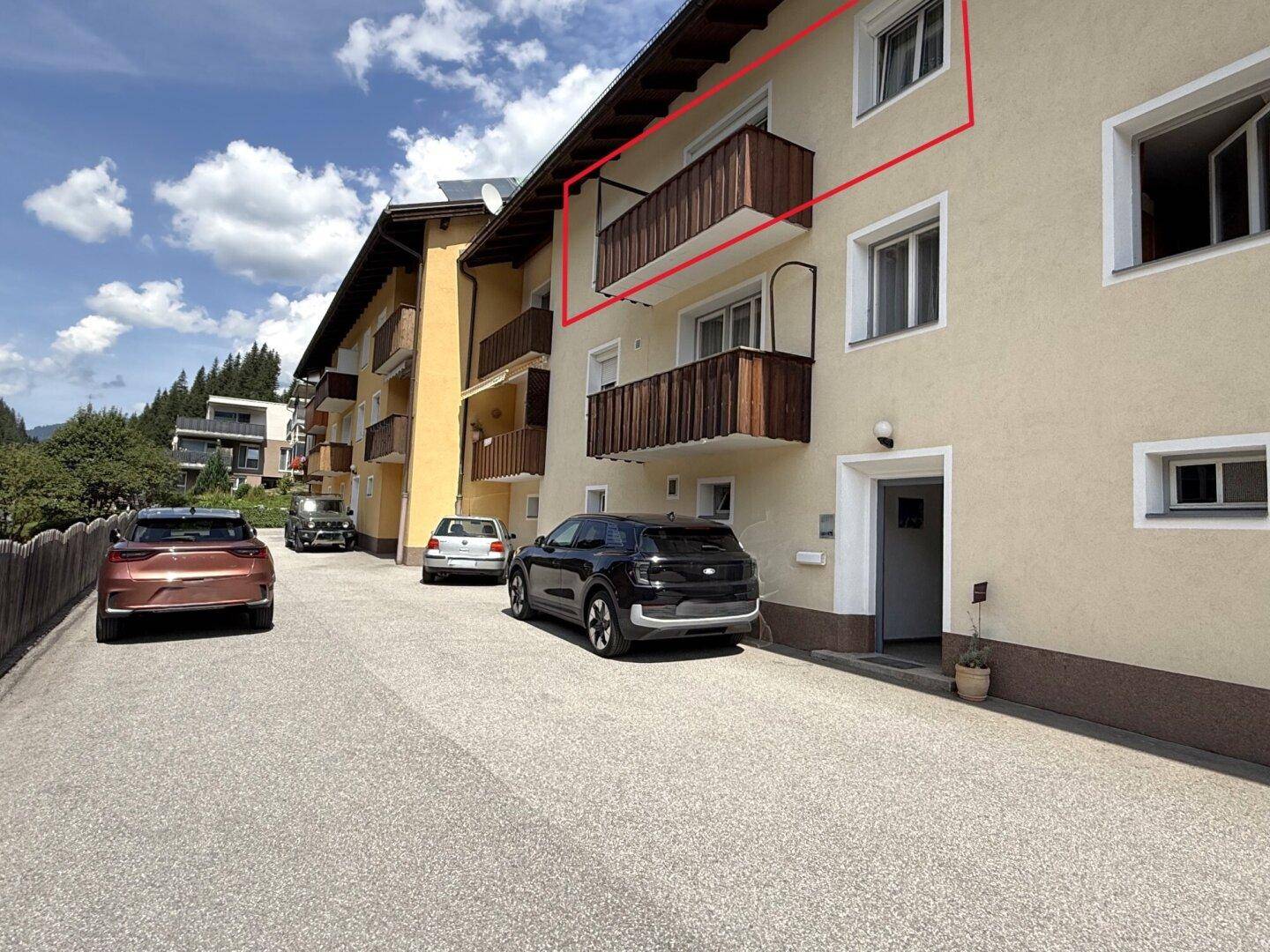 Fassade eines Wohnhauses mit traditionellen Holzbalkonen, Rollläden und gepflasterter Zufahrt mit Parkplätzen.