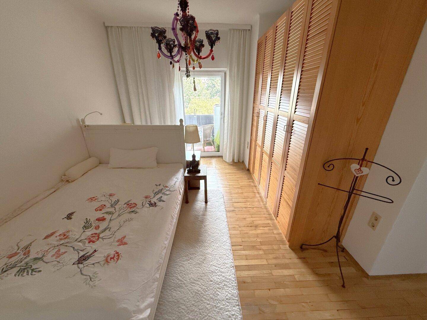 Helles Schlafzimmer mit Einzelbett, Holzschrank und Zugang zum Balkon.