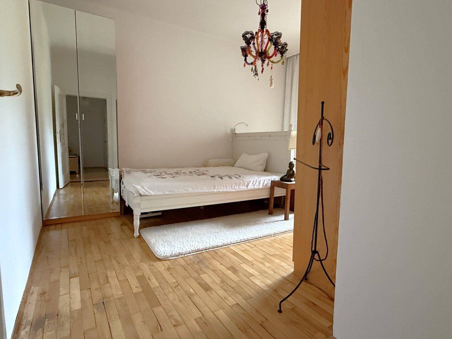 Schlafzimmer mit Holzboden, Bett, Spiegelschrank und farbenfrohem Kronleuchter.