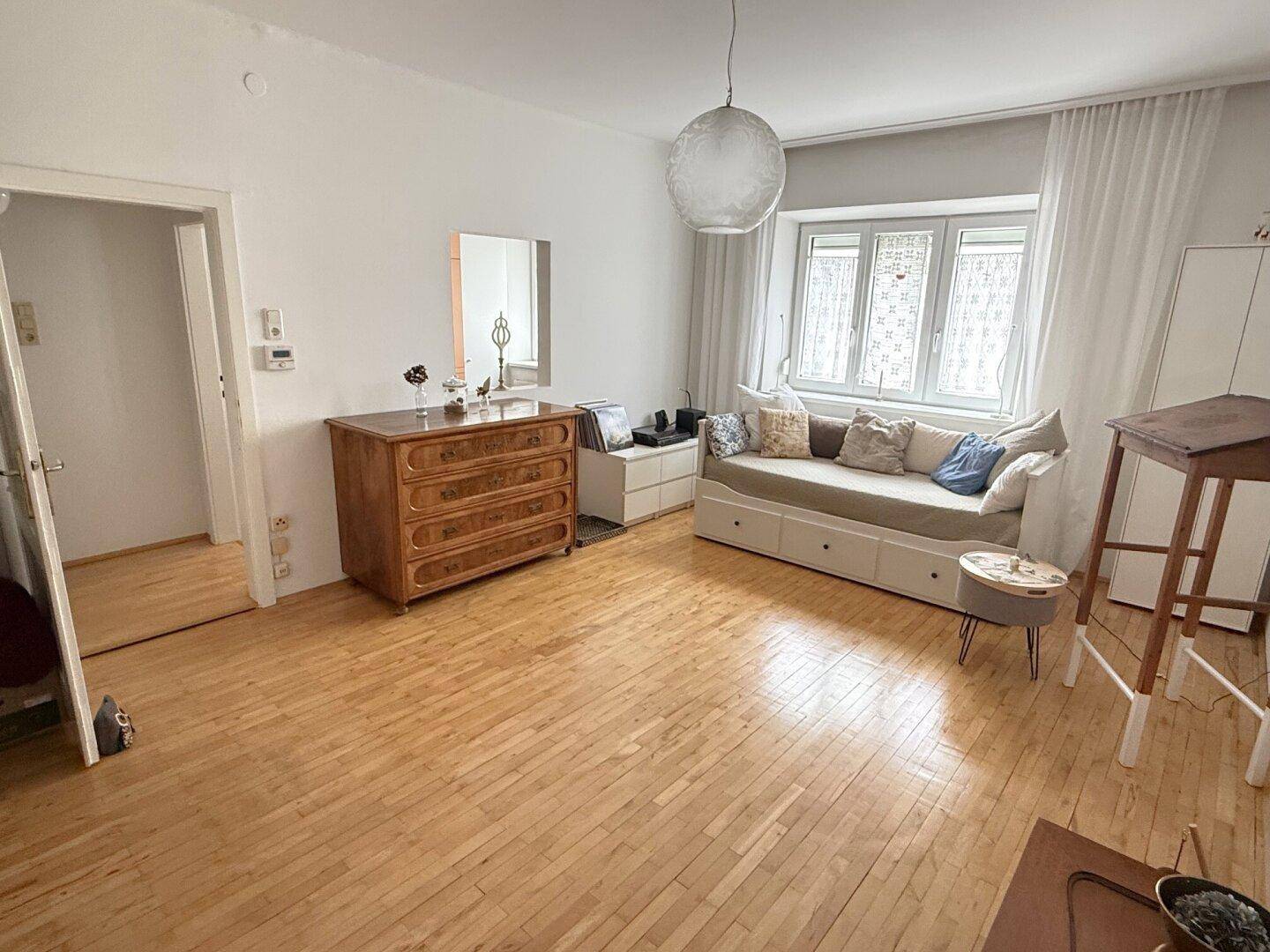 Helles Wohnzimmer mit Holzboden, Tagesbett und klassischer Kommode, mit Zugang zu weiteren Räumen.