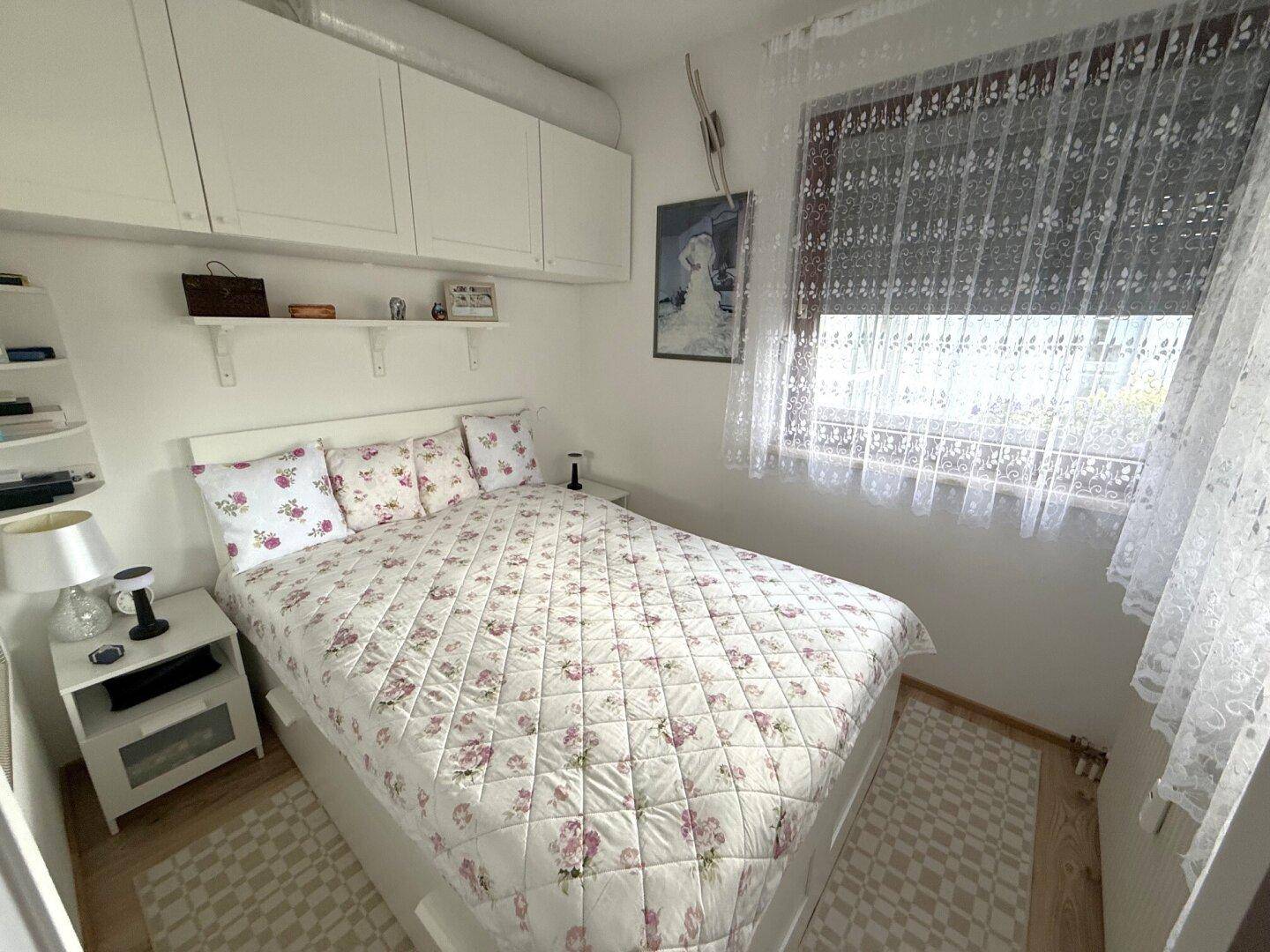 Helles Schlafzimmer mit floraler Bettwäsche, weißen Hängeschränken und Fenster mit Spitzenvorhängen.