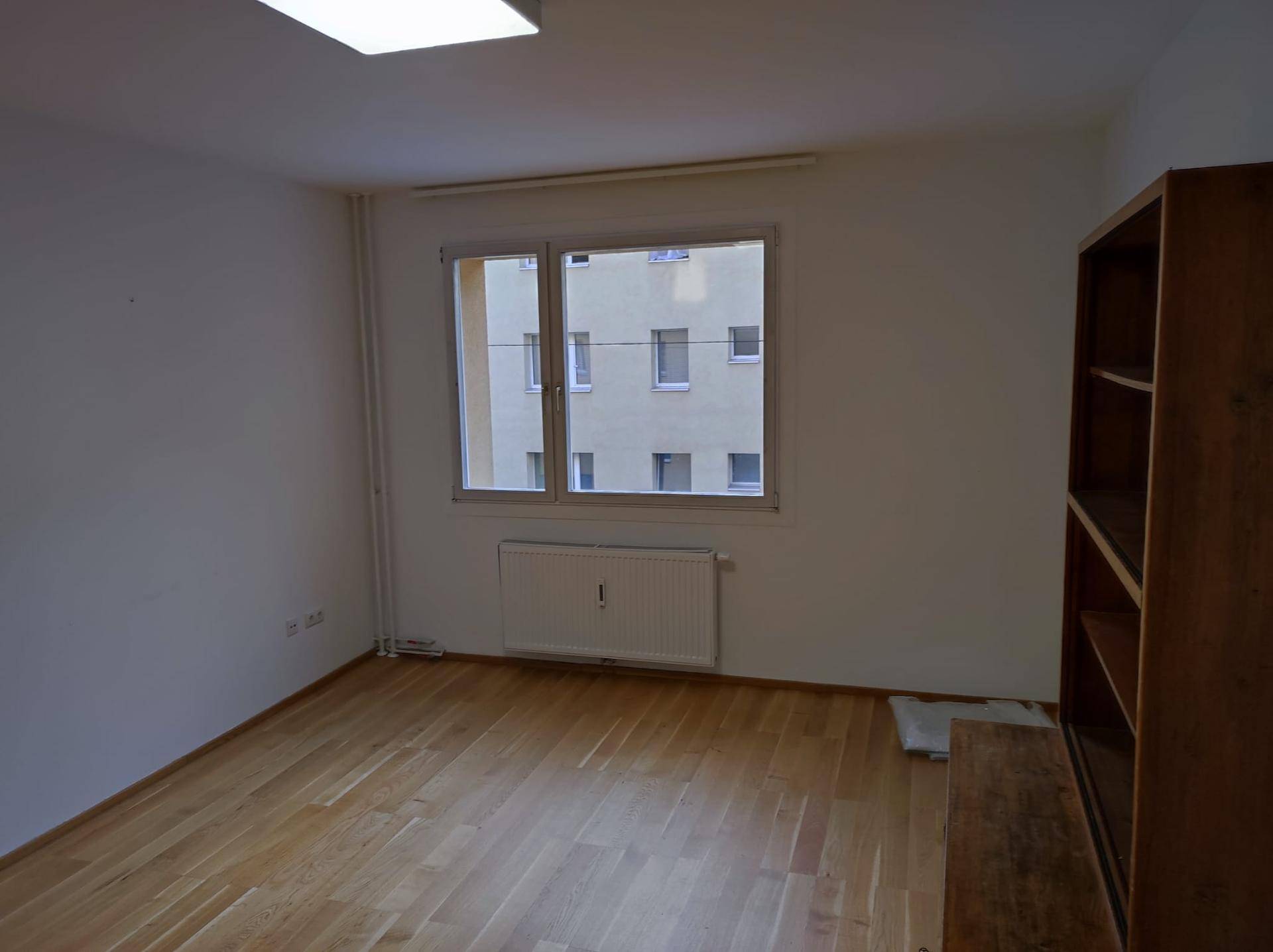Heller Raum mit Parkettboden und einem Fenster, das den Blick auf ein gegenüberliegendes Gebäude freigibt.