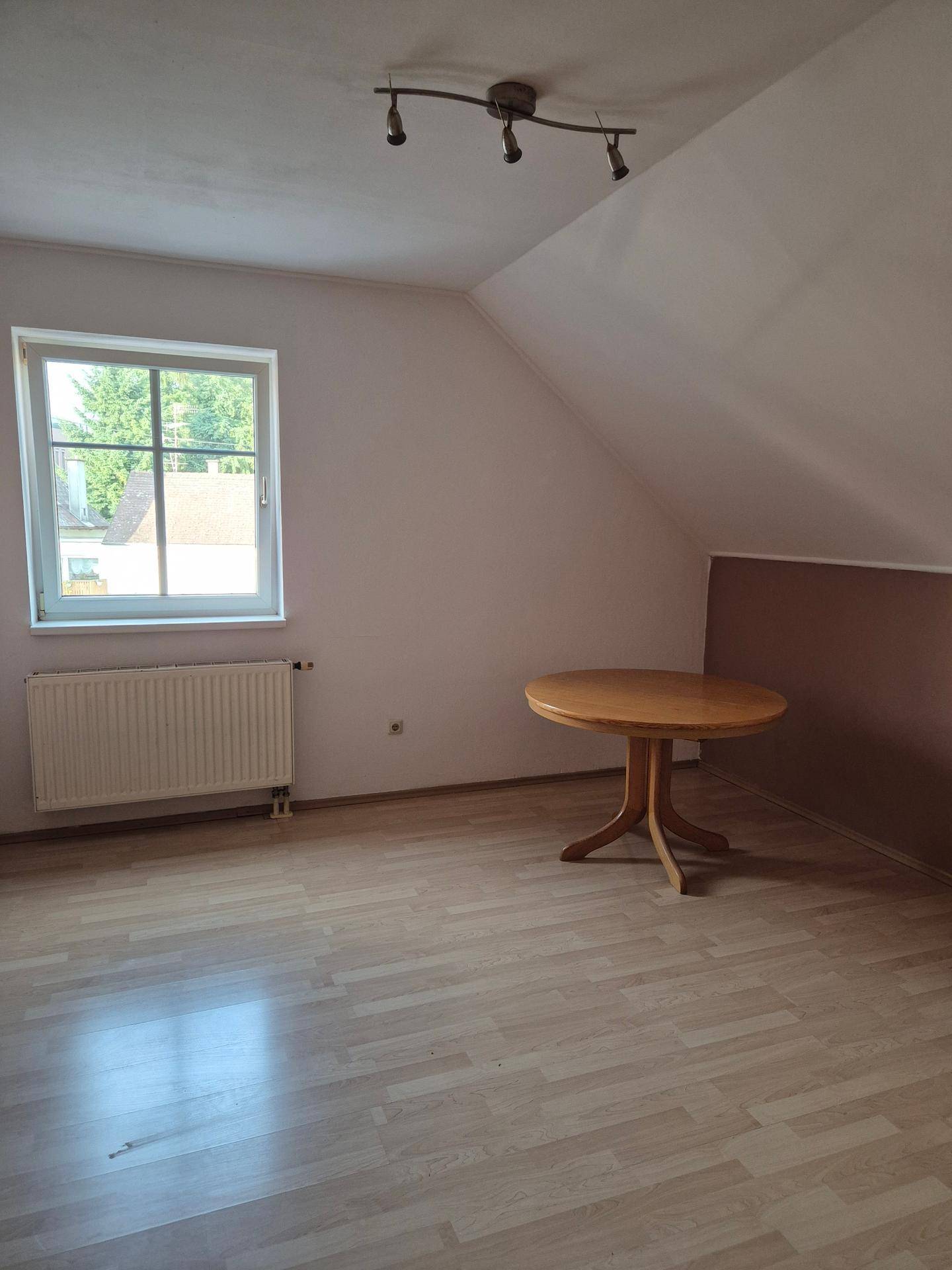 Dachbodenraum mit Dachschrägen, Laminatboden, Fenster, Heizkörper und einem kleinen Holztisch.