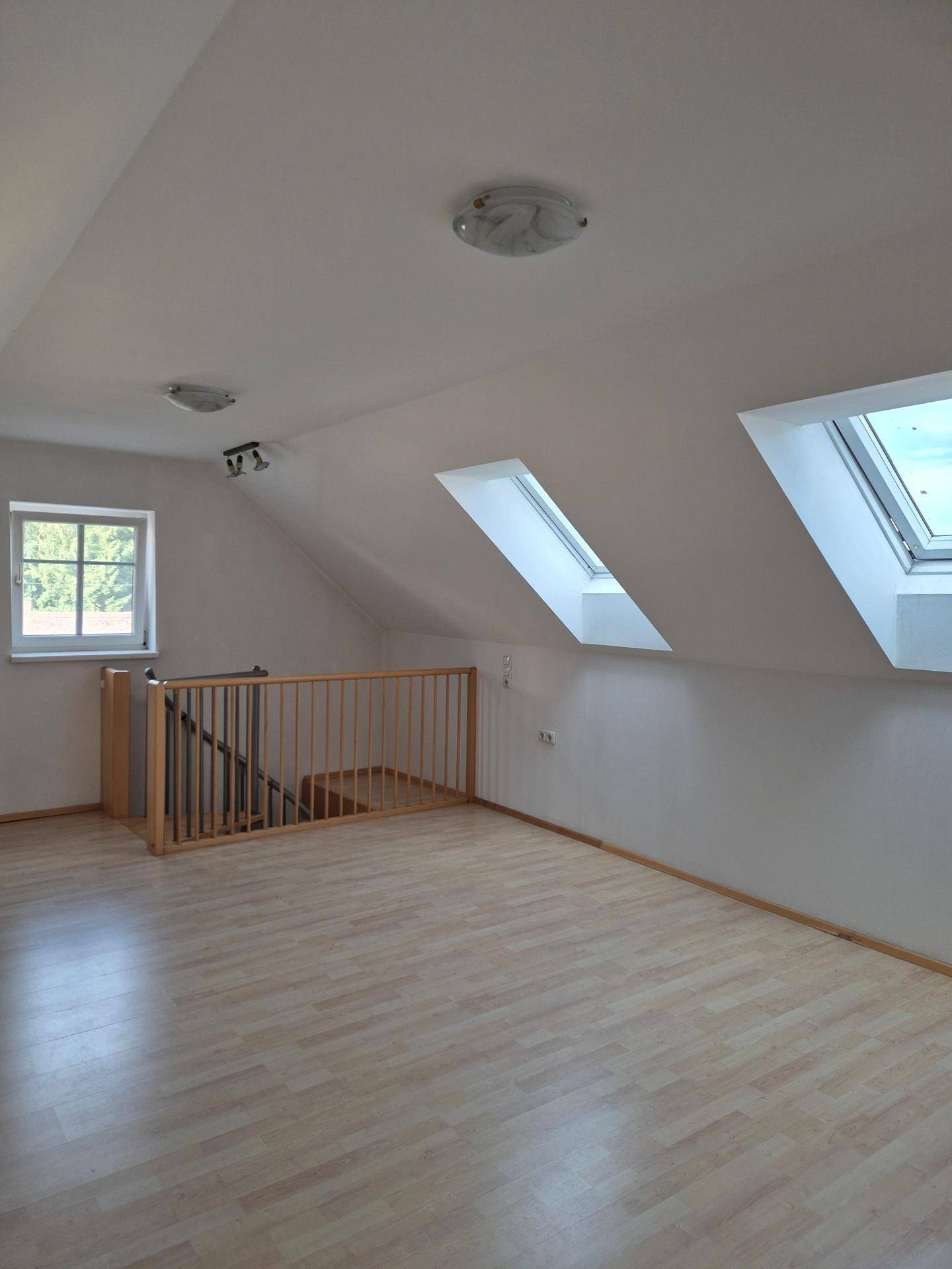 Heller Dachbodenraum mit Dachschrägen, Laminatboden, Holzgeländer und mehreren Dachfenstern.