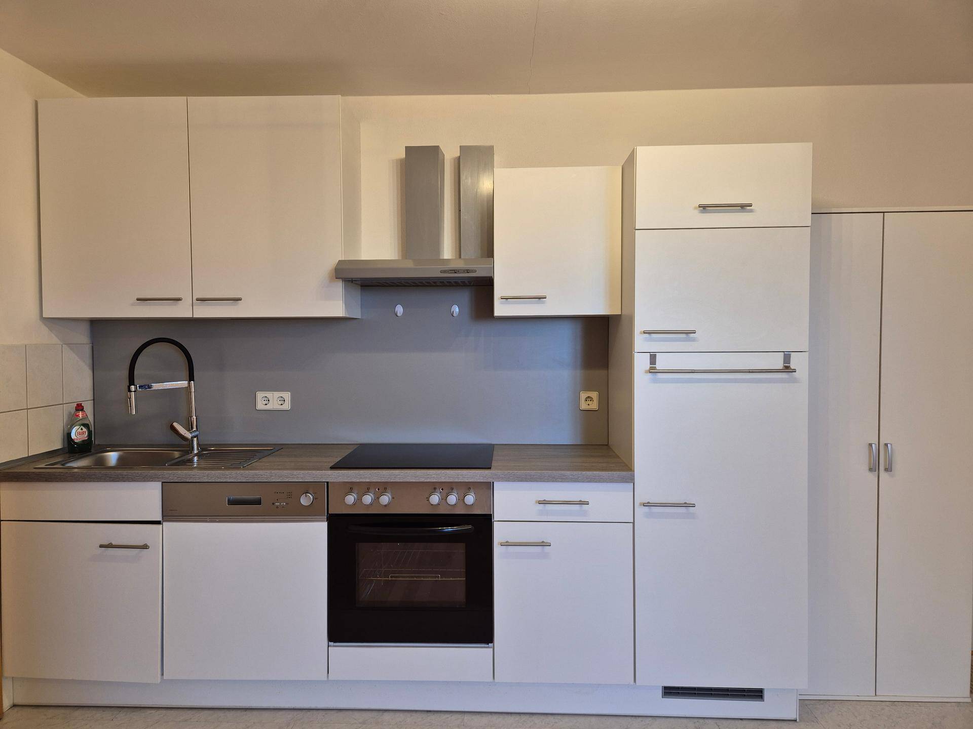 Moderne Einbauküche mit weißen Schränken, integrierten Geräten und grauer Rückwand.