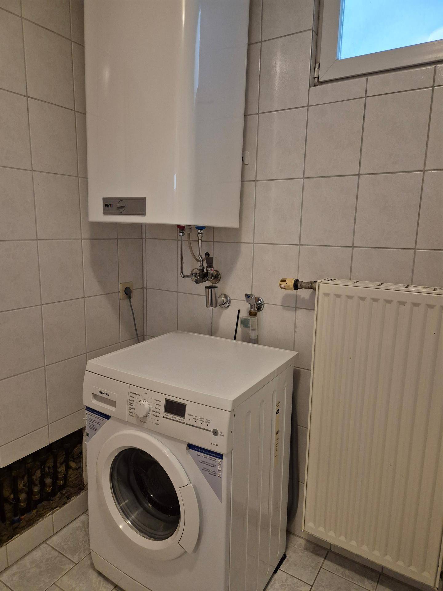 Hauswirtschaftsraum mit Waschmaschine, Warmwasserboiler und Heizkörper an gefliesten Wänden.