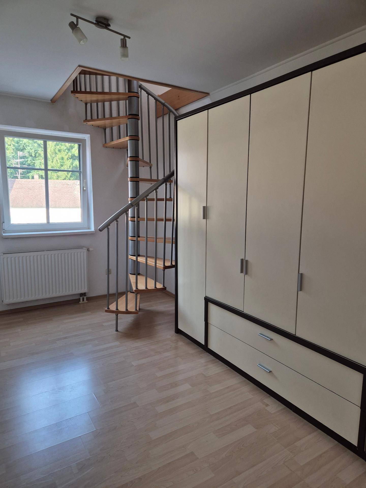 Zimmer mit Laminatboden, Fenster, Heizkörper, Einbauschrank und Wendeltreppe zum Obergeschoss.