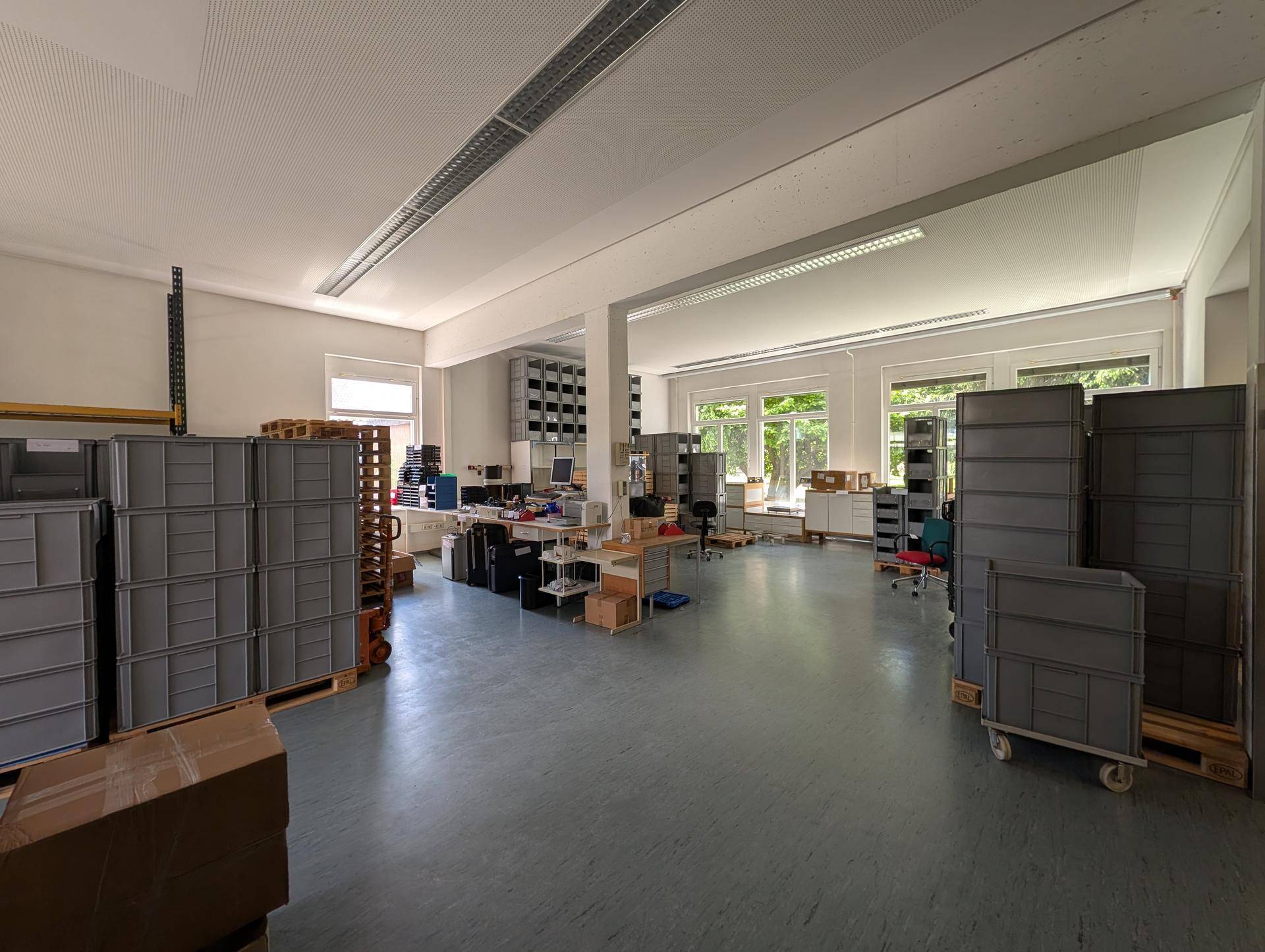 Großer, heller Lagerraum mit vielen Fenstern, Transportboxen und Arbeitsbereichen, ideal für Logistik.