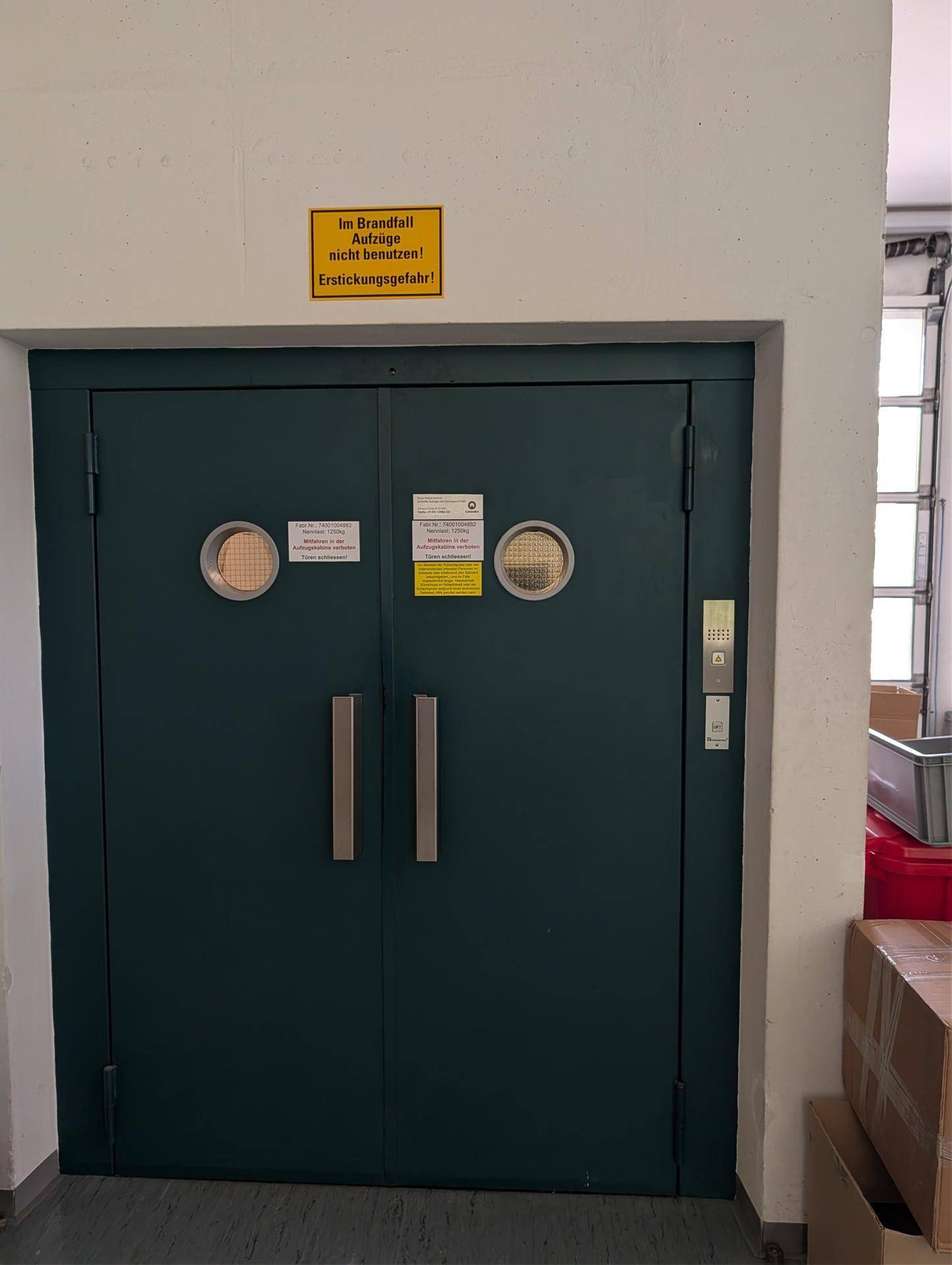 Große, dunkle Doppeltür mit runden Fenstern und einem Warnschild im oberen Bereich des Rahmens.