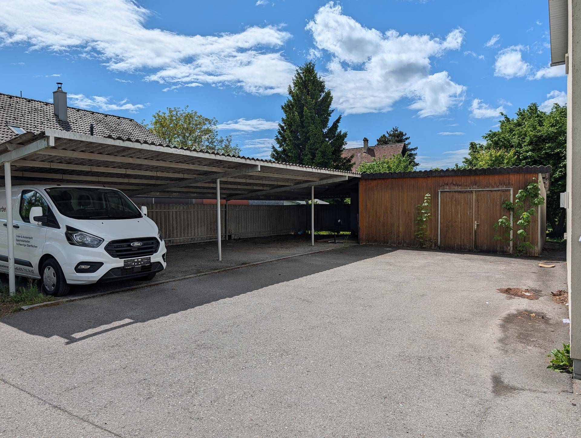 Außenbereich mit einem Carport für Fahrzeuge und einem Holzschuppen im Hintergrund unter blauem Himmel.