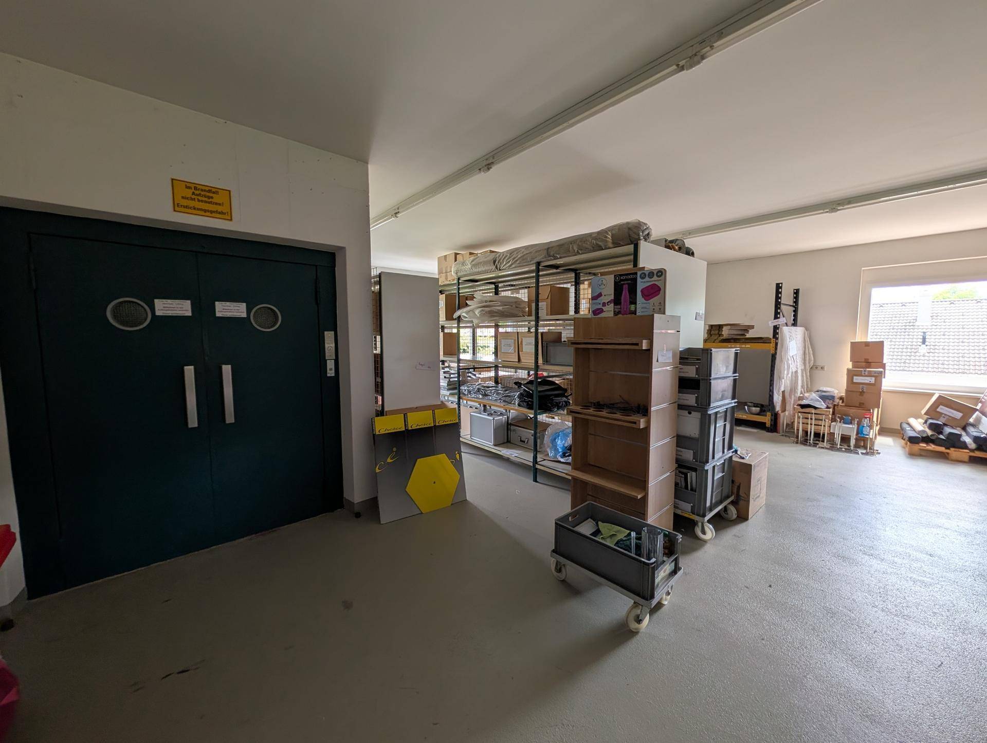 Großer Lagerraum mit Industrietüren, Betonboden und Regalen, hell durch ein Fenster im Hintergrund.