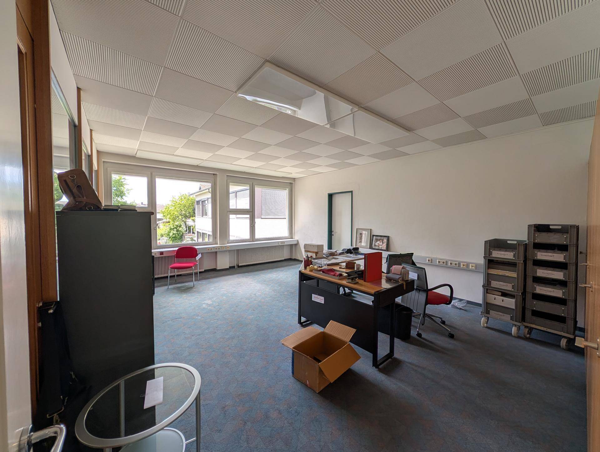 Helles Büro mit Teppichboden, großen Fenstern und einem Schreibtisch in der Mitte des Raumes.