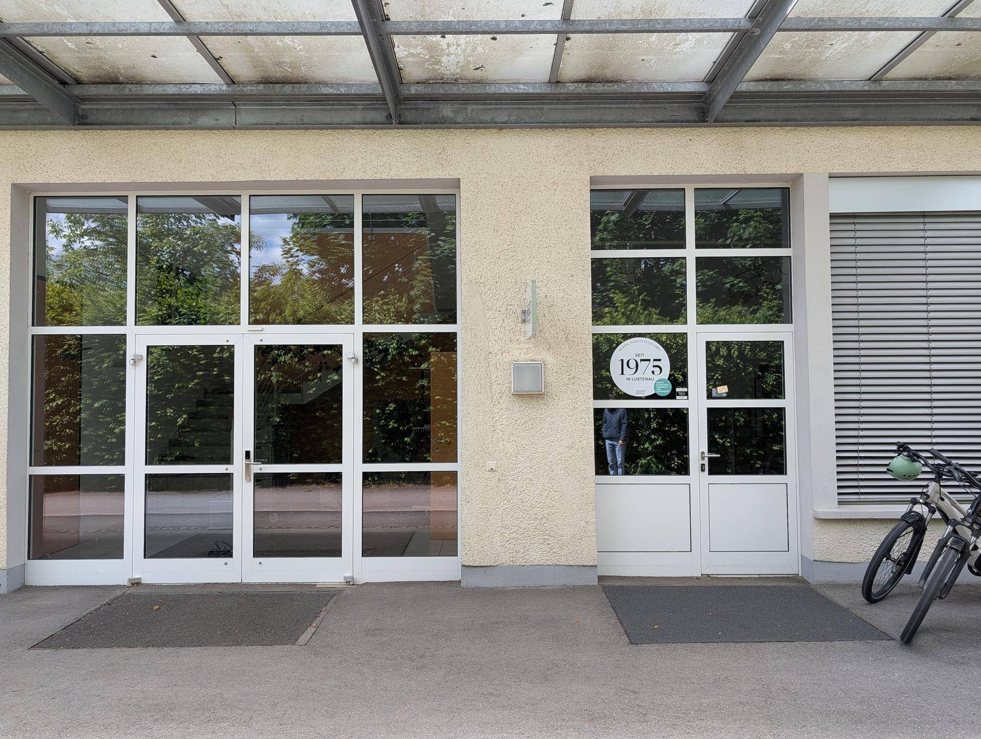 Eingangsbereich eines modernen Gebäudes mit großen Glasfenstern und einer Tür mit Firmenlogo.