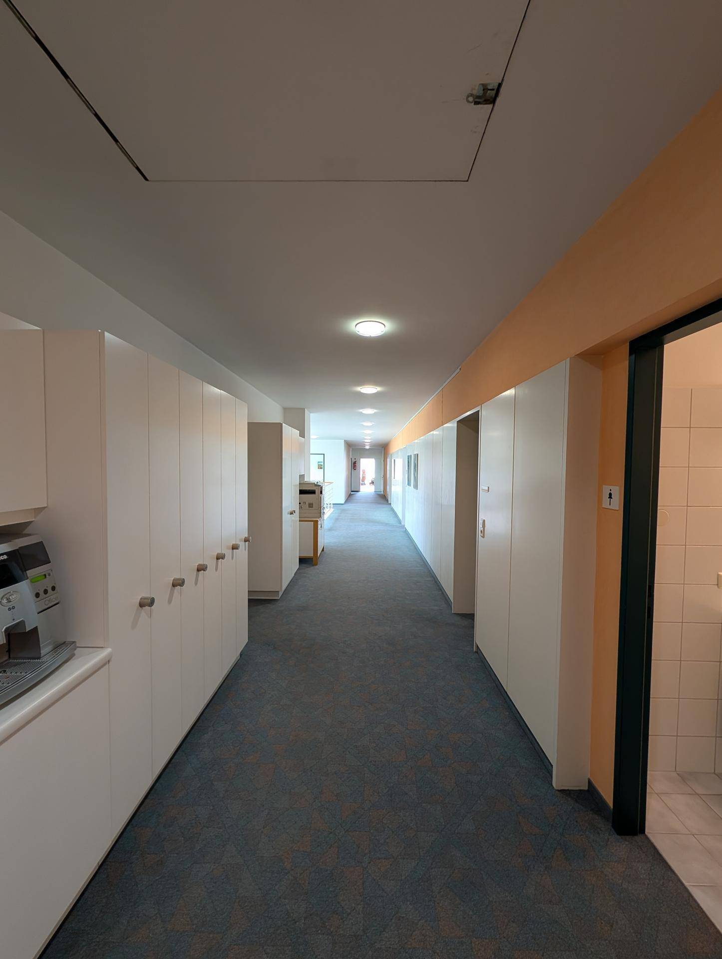 Langer Bürogang mit vielen weißen Schränken und Zugang zu den Toiletten, funktional und hell.