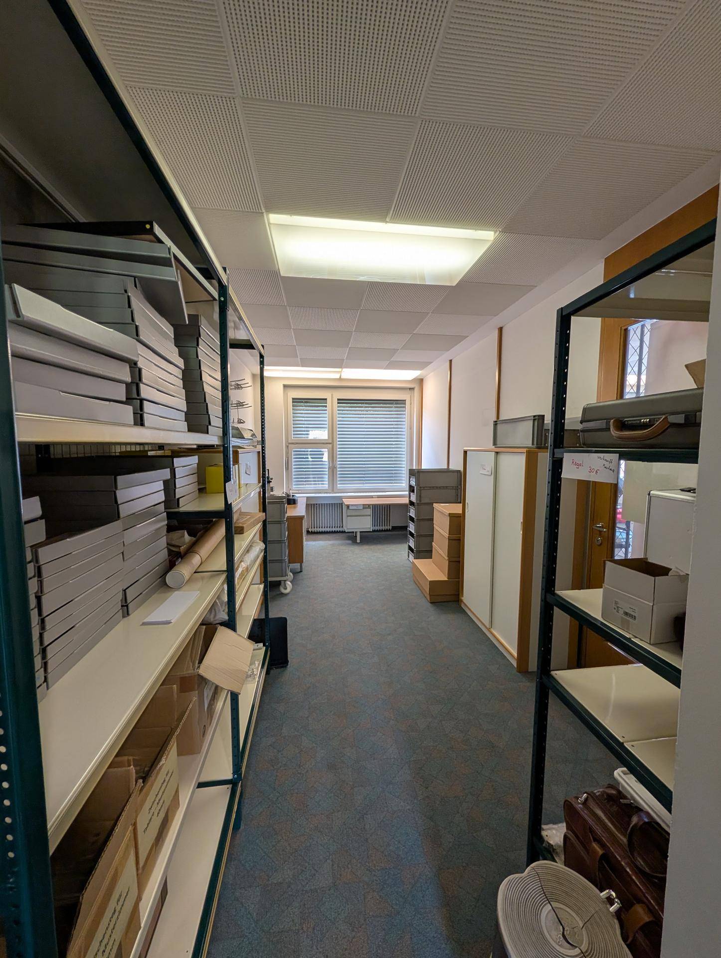 Langer Lagerraum mit Regalen, Teppichboden und einem Fenster am Ende, ideal für Büromaterial.