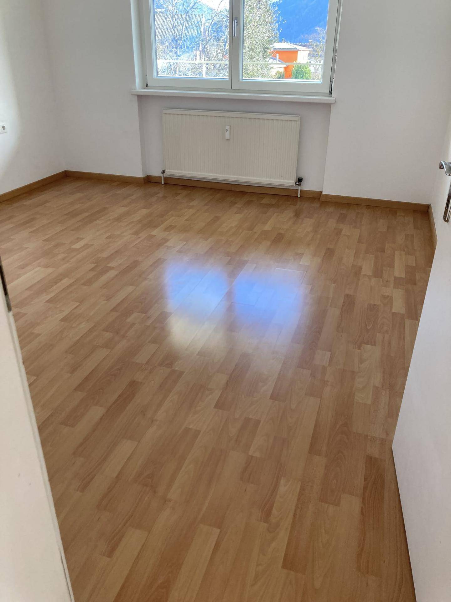 Helles, leeres Zimmer mit Laminatboden und einem Fenster mit Blick ins Grüne.