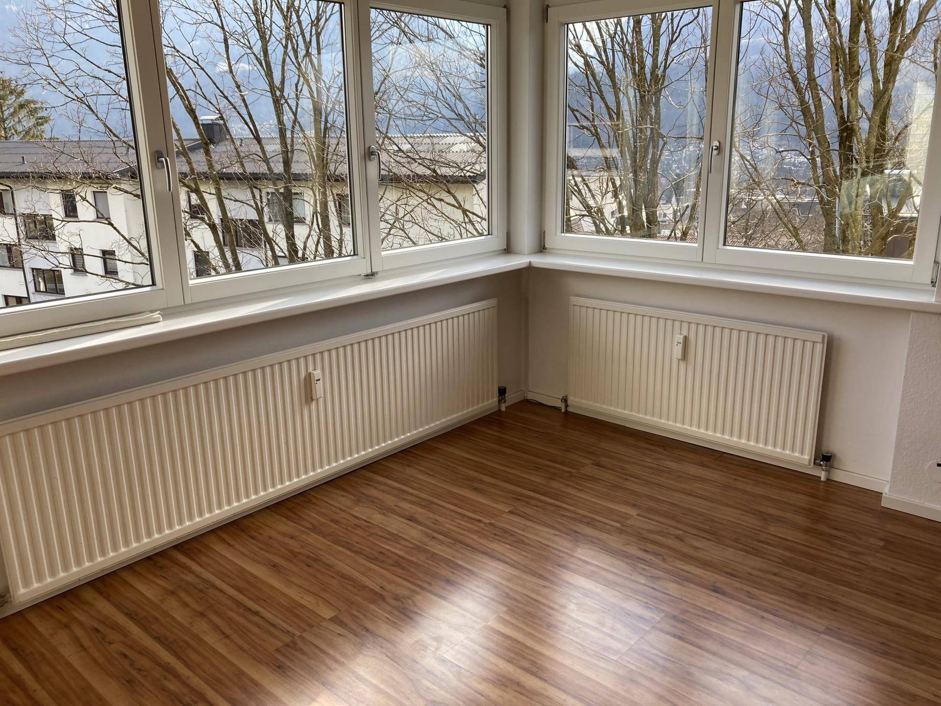 Lichtdurchfluteter Raum mit großen Fenstern, Laminatboden und Heizkörpern unter den Fenstern.