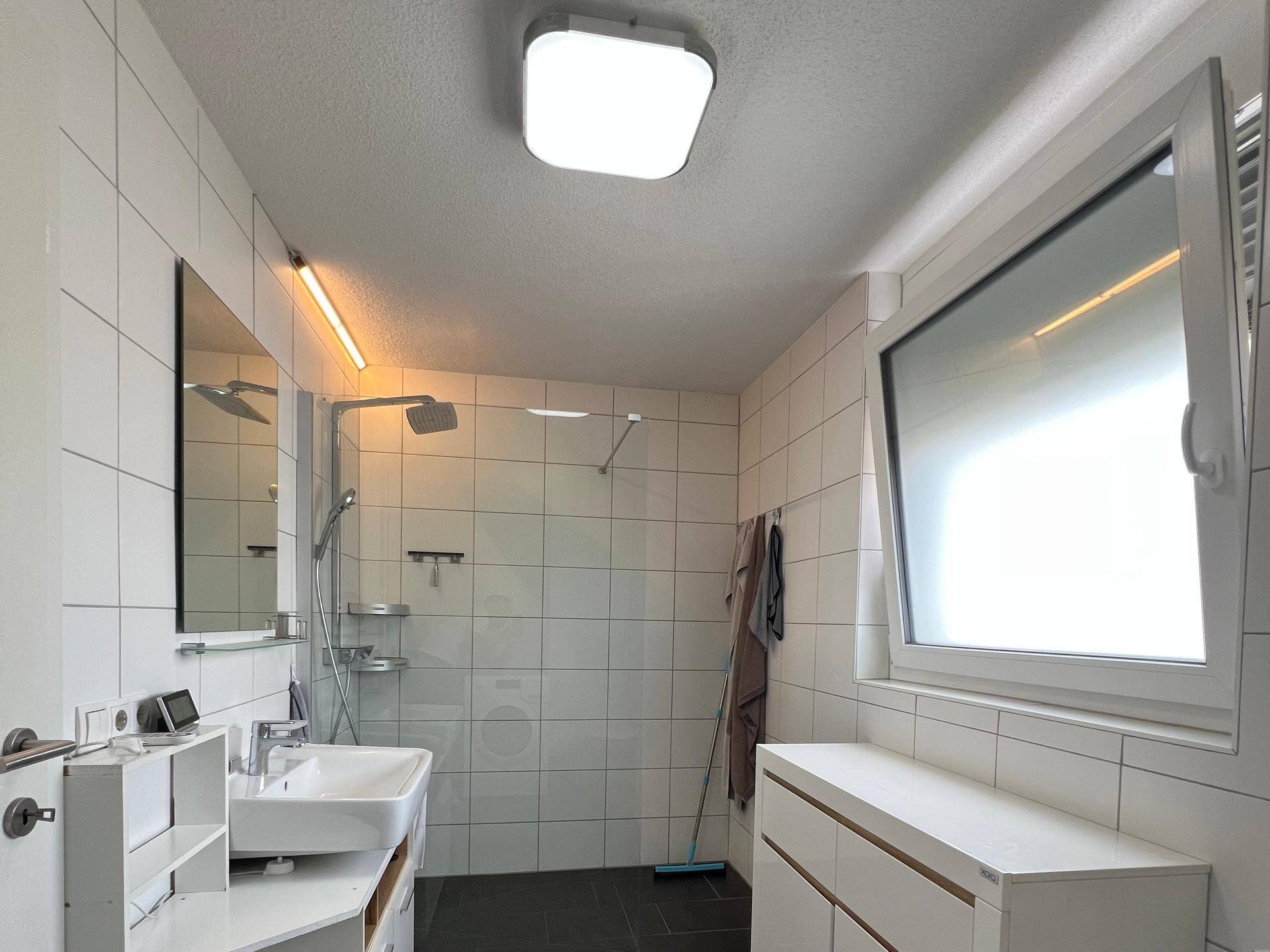 Modernes Badezimmer mit großer Dusche, weißen Fliesen und Fenster für Tageslicht.
