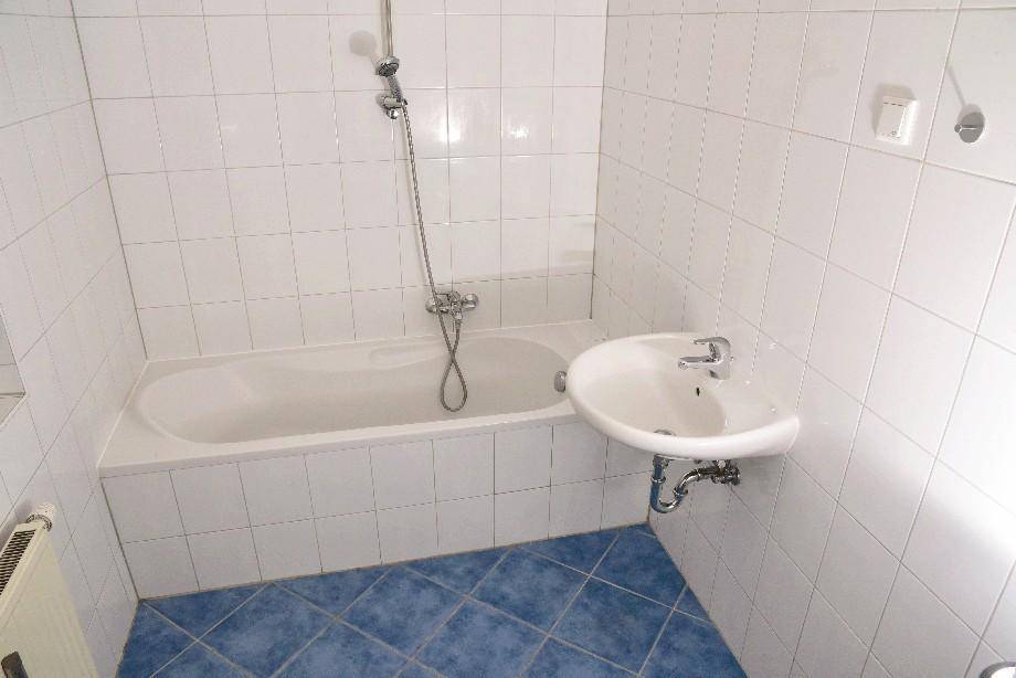 Badezimmer mit weißen Wandfliesen, blauen Bodenfliesen, Badewanne mit Duschfunktion und Waschbecken.