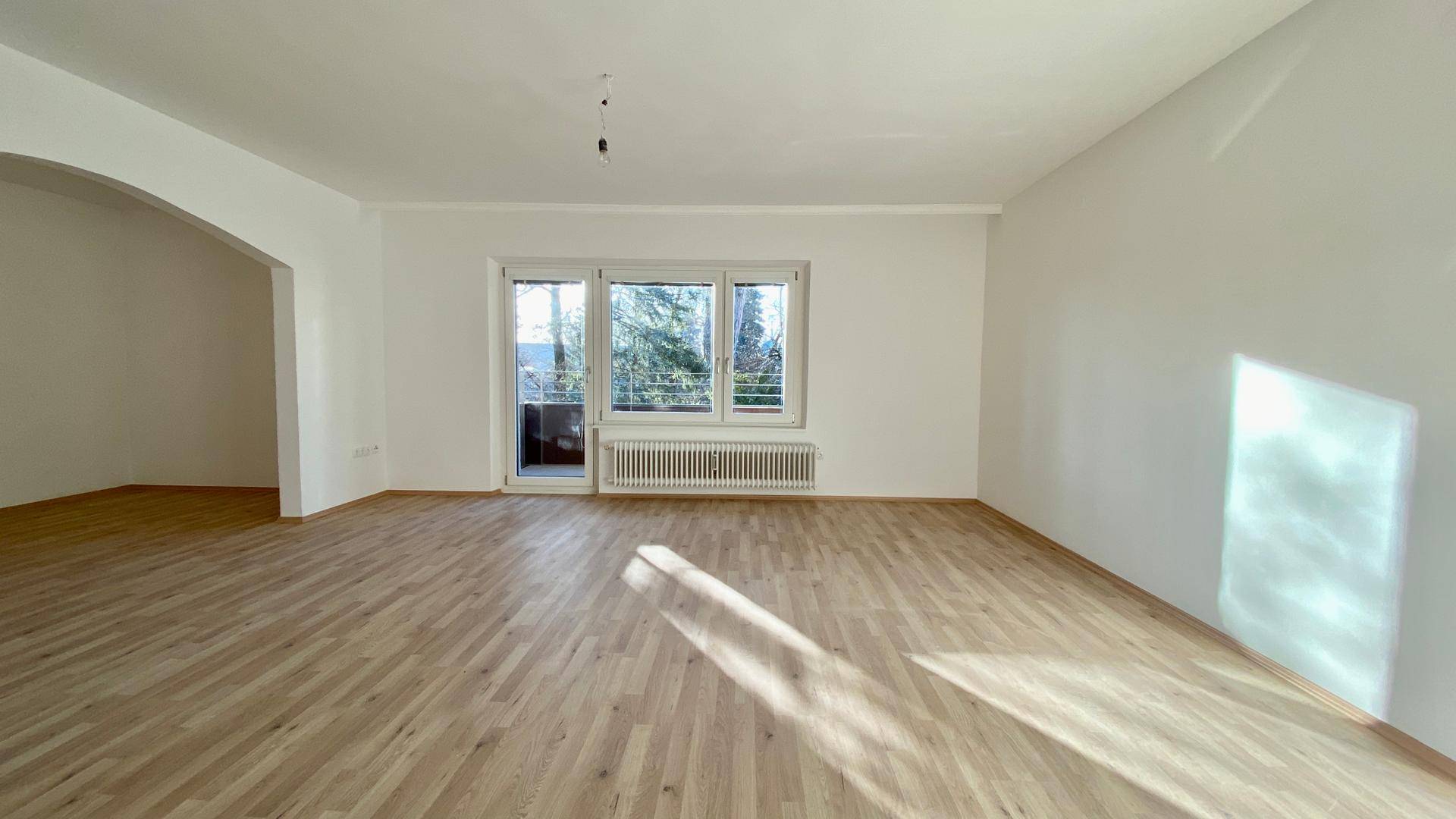 Geräumiges Wohnzimmer mit hellem Parkettboden und großen Fenstern, die viel Licht hereinlassen.