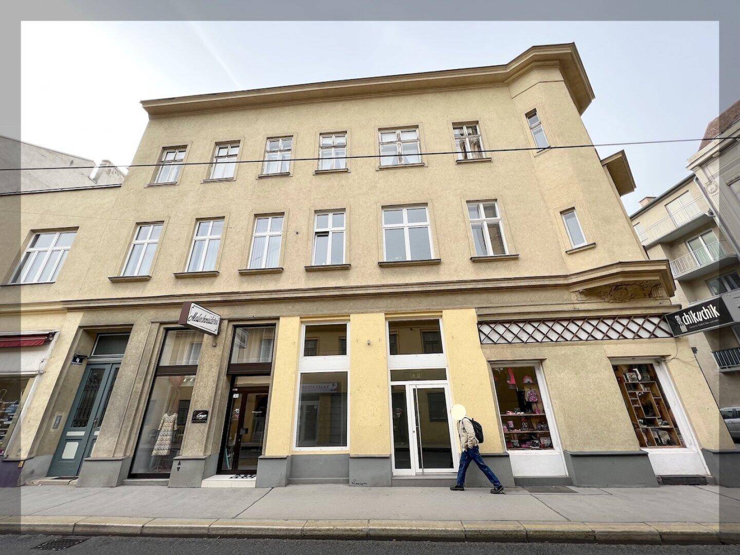Gelbes Eckgebäude mit großen Schaufenstern im Erdgeschoss und Wohnungen darüber an einer Straße.