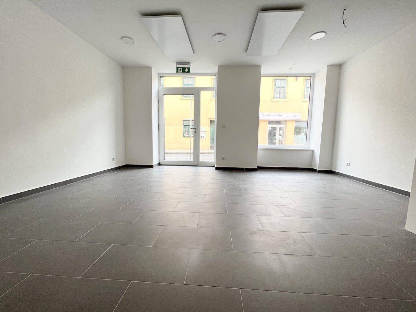 Geräumiger Gewerberaum mit großen Schaufenstern, modernen Deckenleuchten und Fliesenboden.