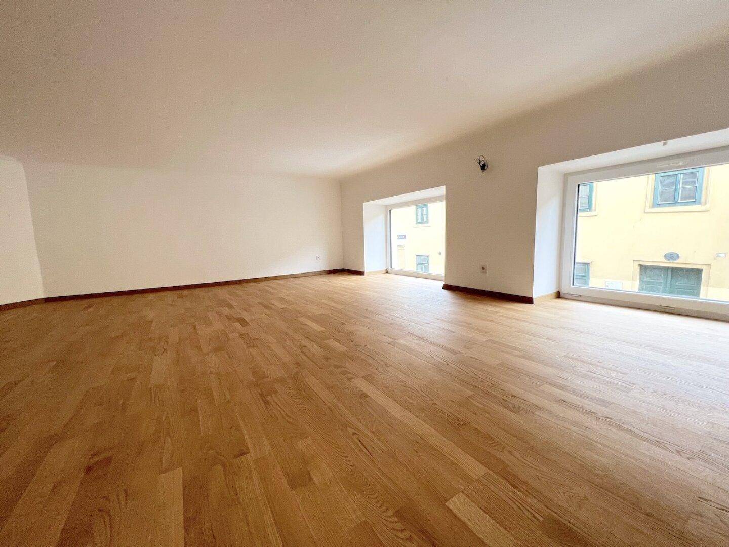 Großer, heller Raum mit Holzboden und breiten Fenstern, die einen Blick auf die Stadt bieten.