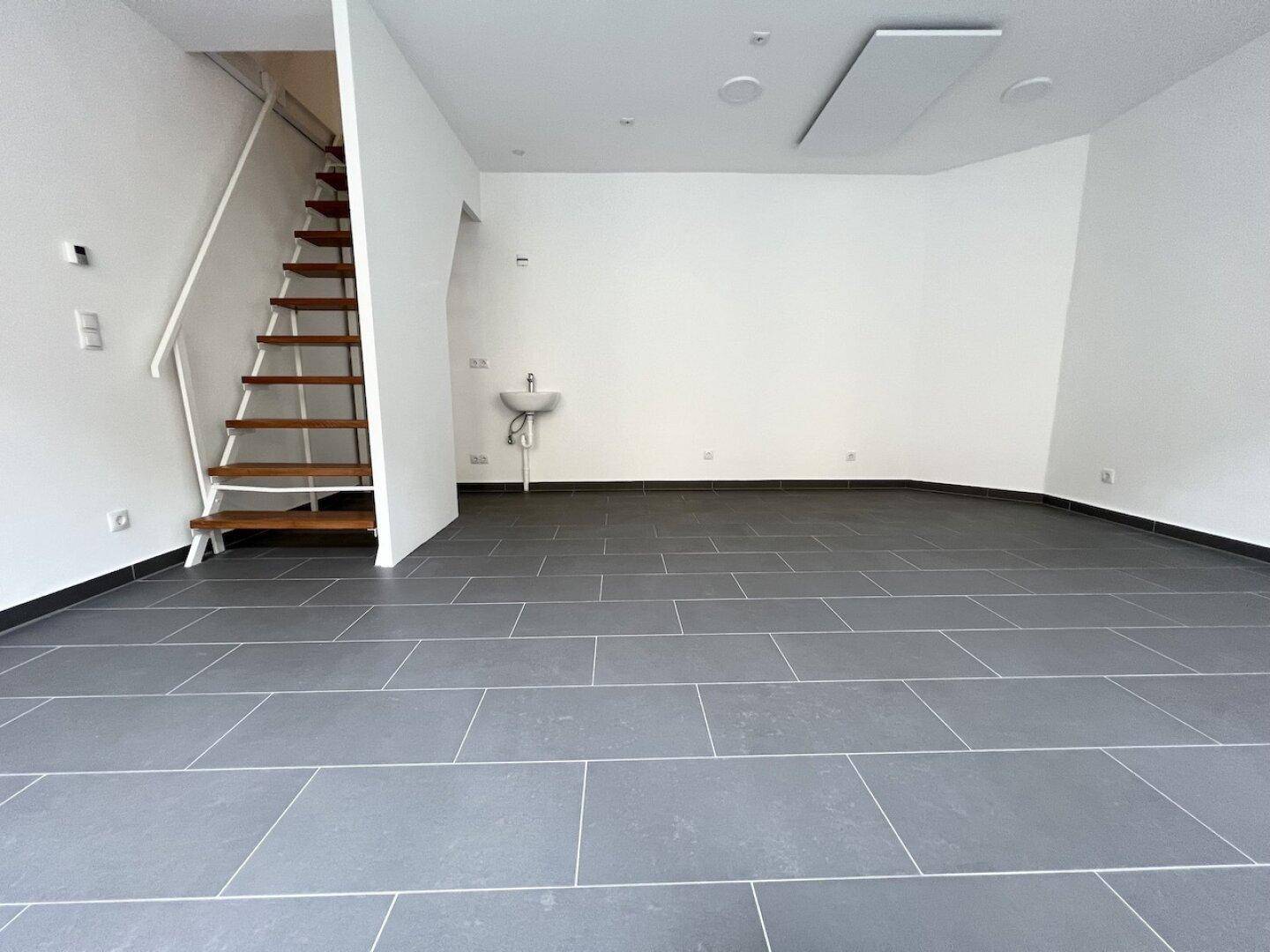 Heller Gewerberaum mit Holztreppe, Waschbecken und dunklem Fliesenboden in neuwertigem Zustand.