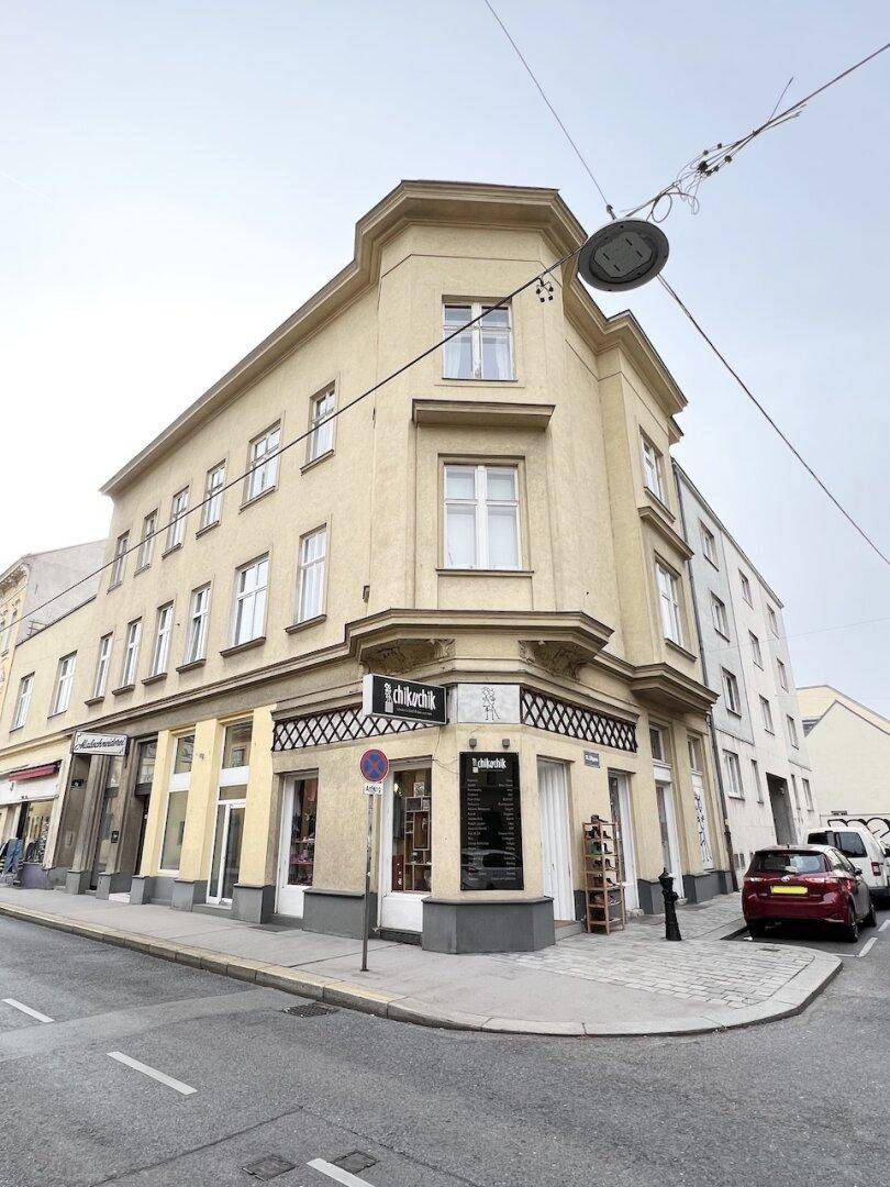 Historisches Eckgebäude an einer belebten Straße mit Geschäften im Erdgeschoss und parkenden Autos.