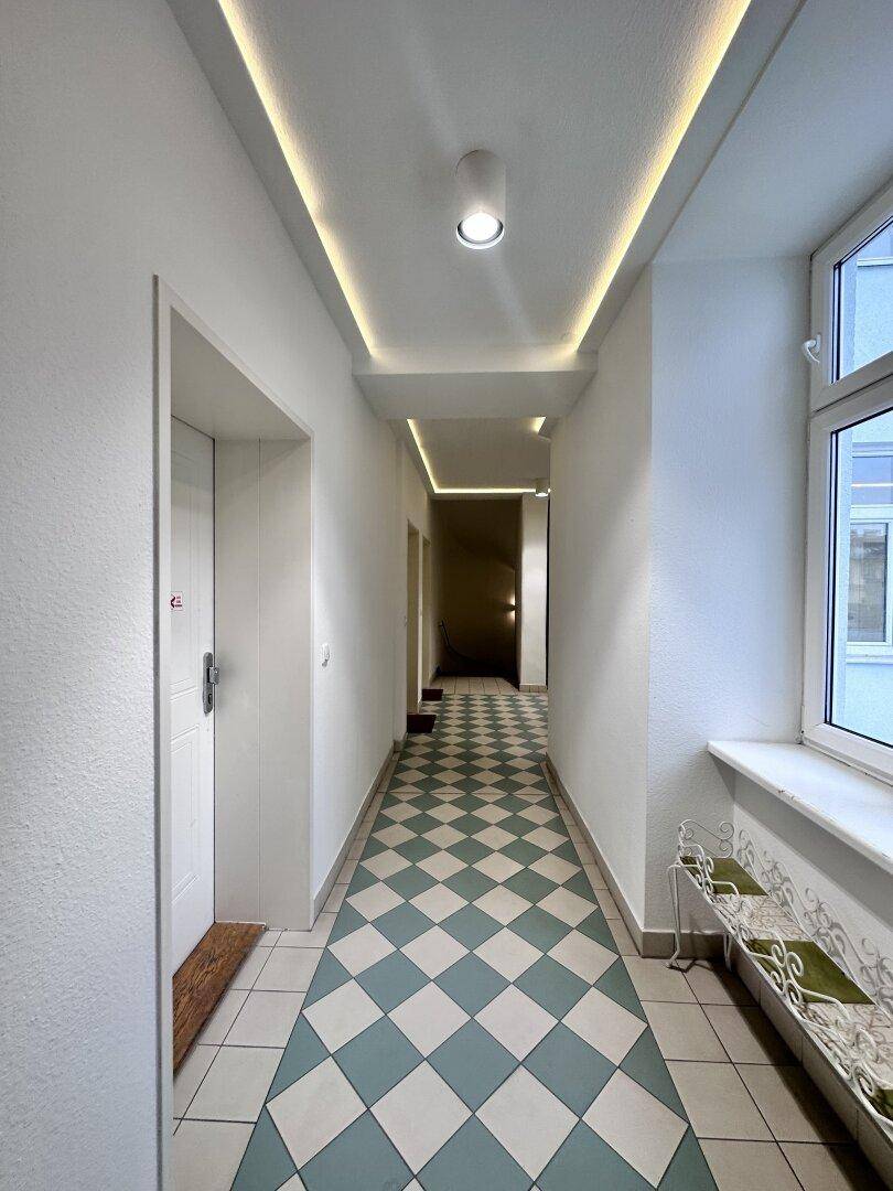 Langer Flur mit Schachbrettmuster-Fliesenboden und Fenster für natürliches Licht.