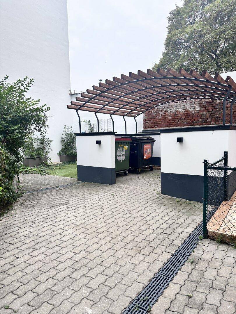 Gepflasterter Außenbereich mit Mülltonnen und einer Holzpergola.