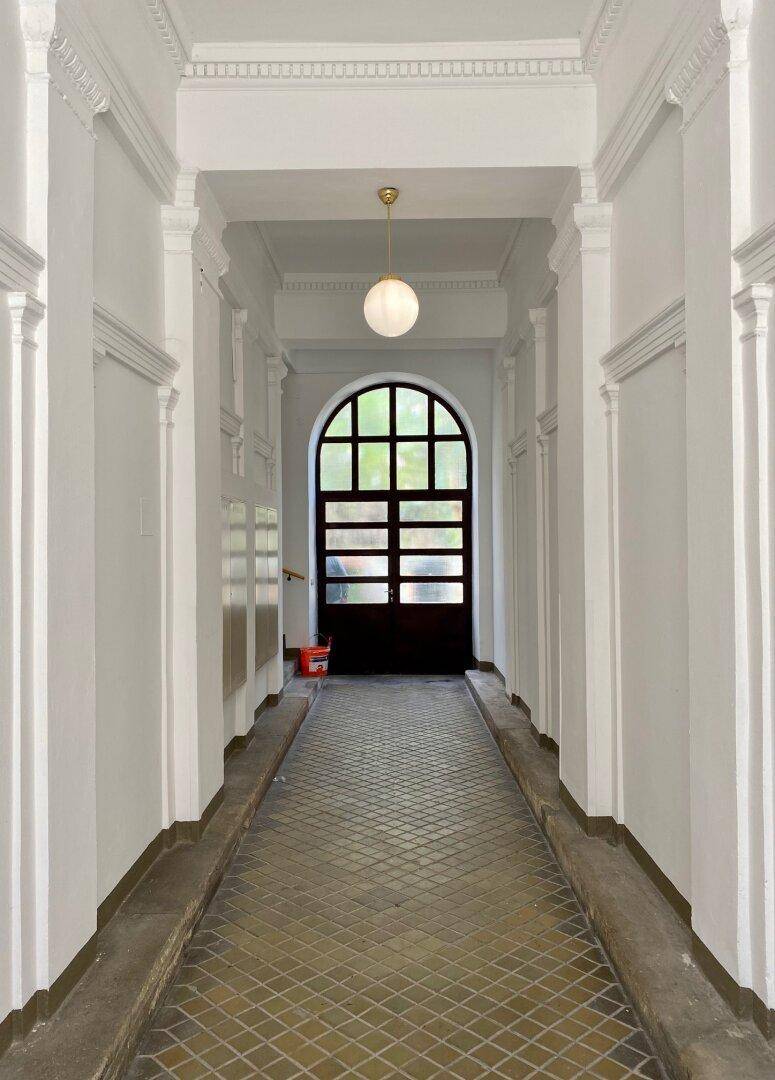 Historischer Hausflur mit hohen Decken, Stuckelementen und einem Bogenfenster am Ende.
