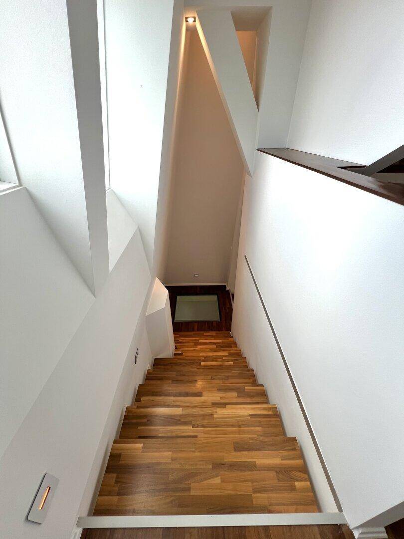 Moderne Holztreppe führt nach unten, mit hellen Wänden und indirekter Beleuchtung.