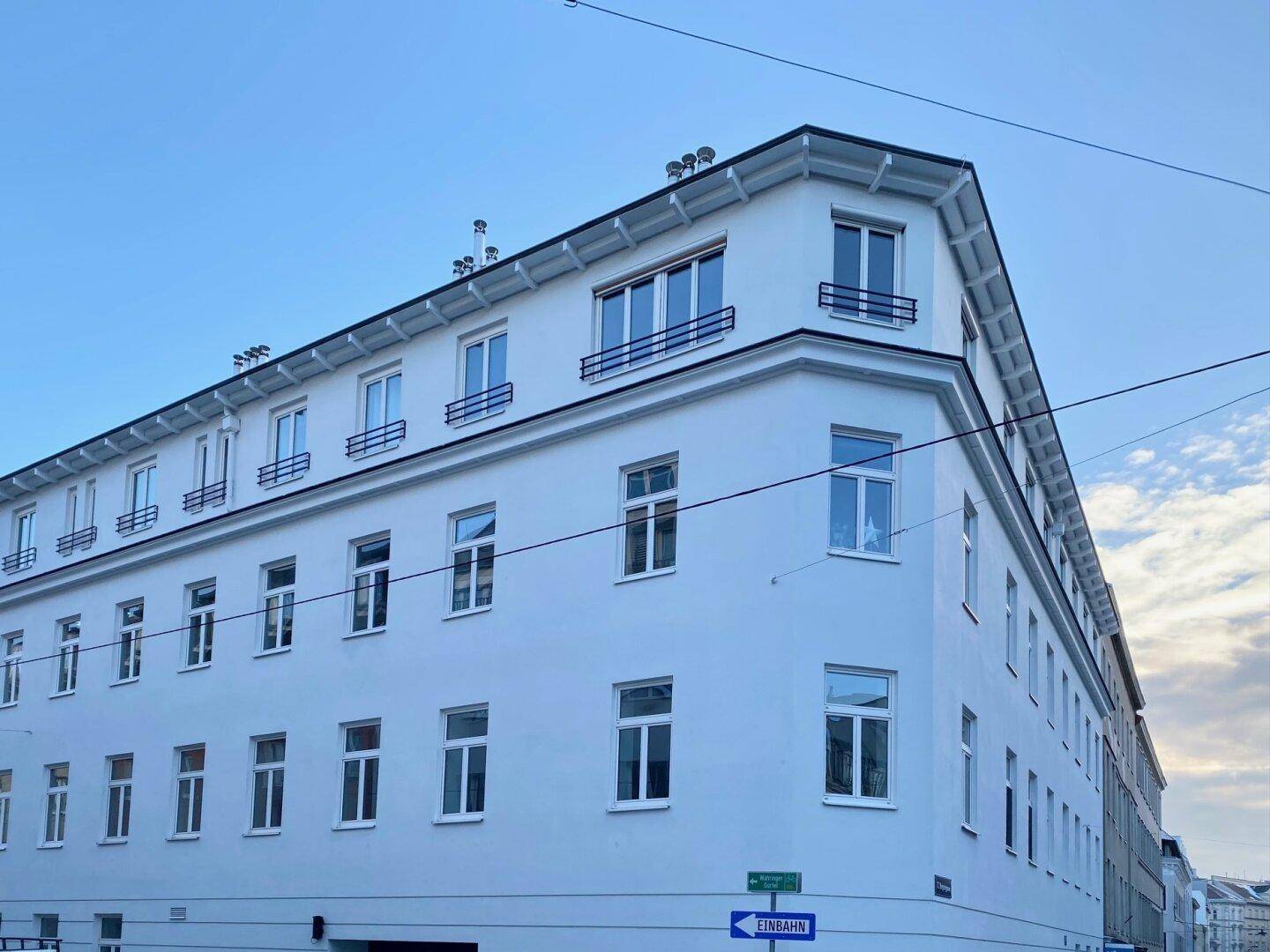 Weißes Eckgebäude mit vielen Fenstern und einem Mansarddach unter klarem Himmel.