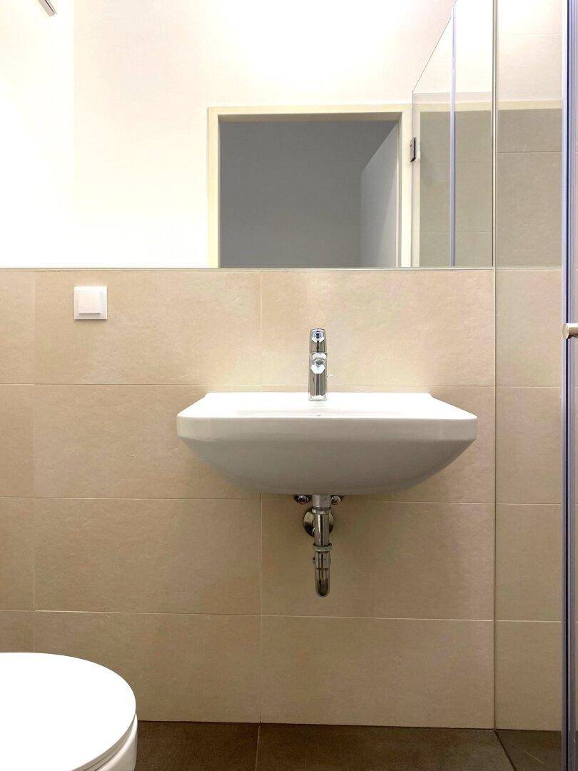 Nahaufnahme des Badezimmers mit Einzelwaschbecken, Spiegel und beigefarbenen Wandfliesen, inklusive Toilettenausschnitt.