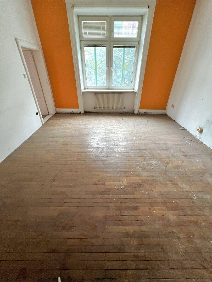 Leerer Raum mit orangefarbener Akzentwand, Holzdielenboden und einem Fenster mit Heizkörper darunter.