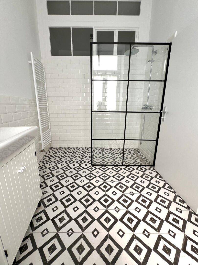 Helles Badezimmer mit moderner Duschkabine, Fenster und geometrischem Fliesenboden.