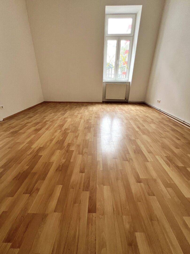 Helles Zimmer mit Parkettboden, schräger Wand und einem hohen Fenster mit Blick nach draußen.