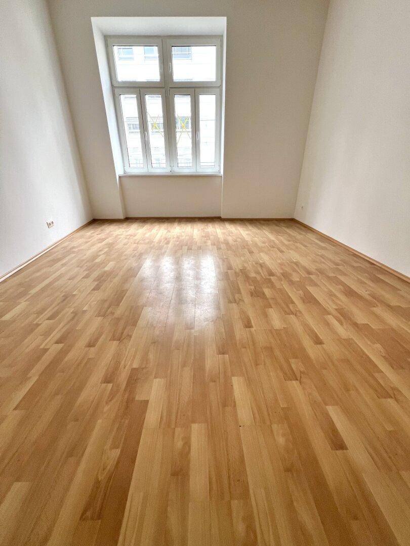 Großes, helles Zimmer mit Parkettboden und einem breiten Fenster, das viel Licht hereinlässt.