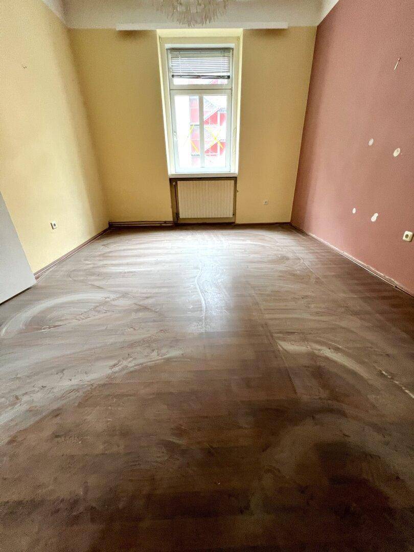Leerer Raum mit Holzboden, Fenster und Wänden in verschiedenen Farbtönen.