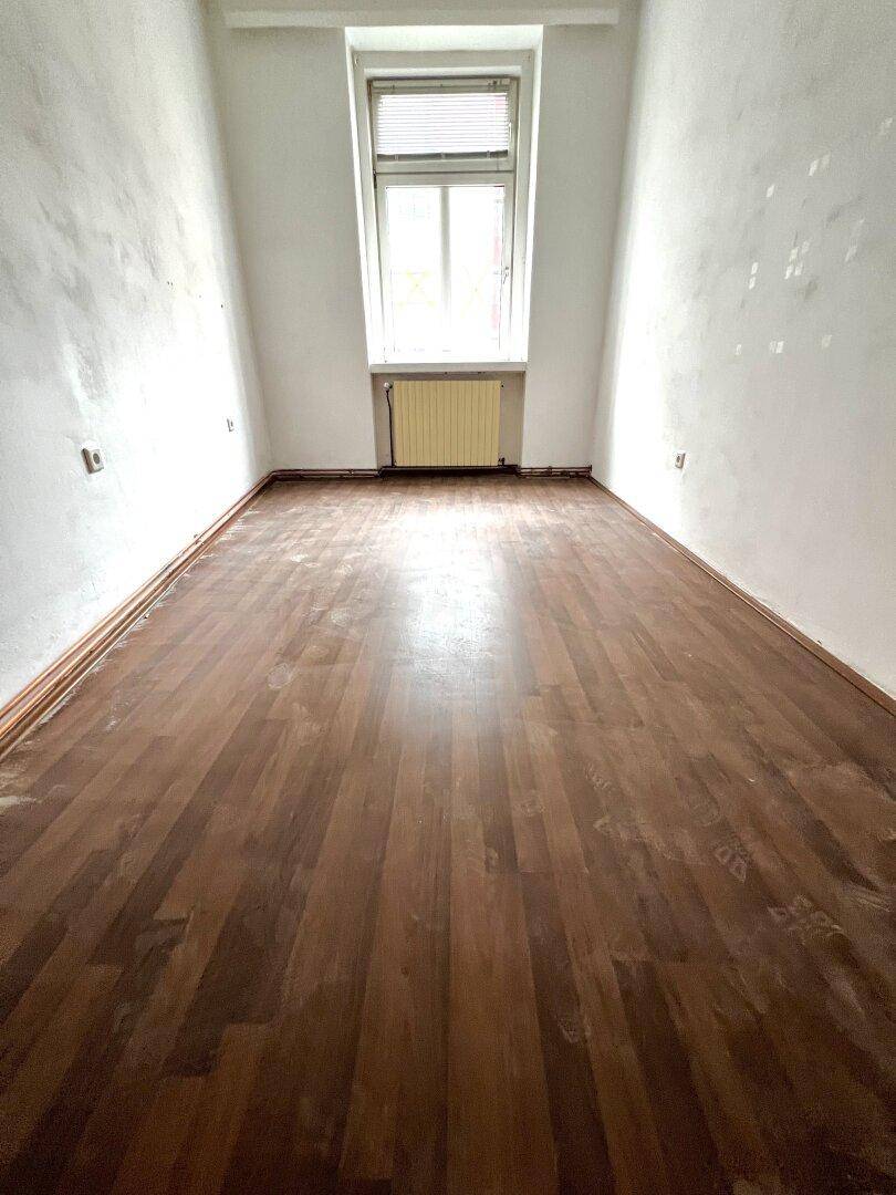 Leerer Raum mit Holzboden und einem Fenster, das Tageslicht hereinlässt.