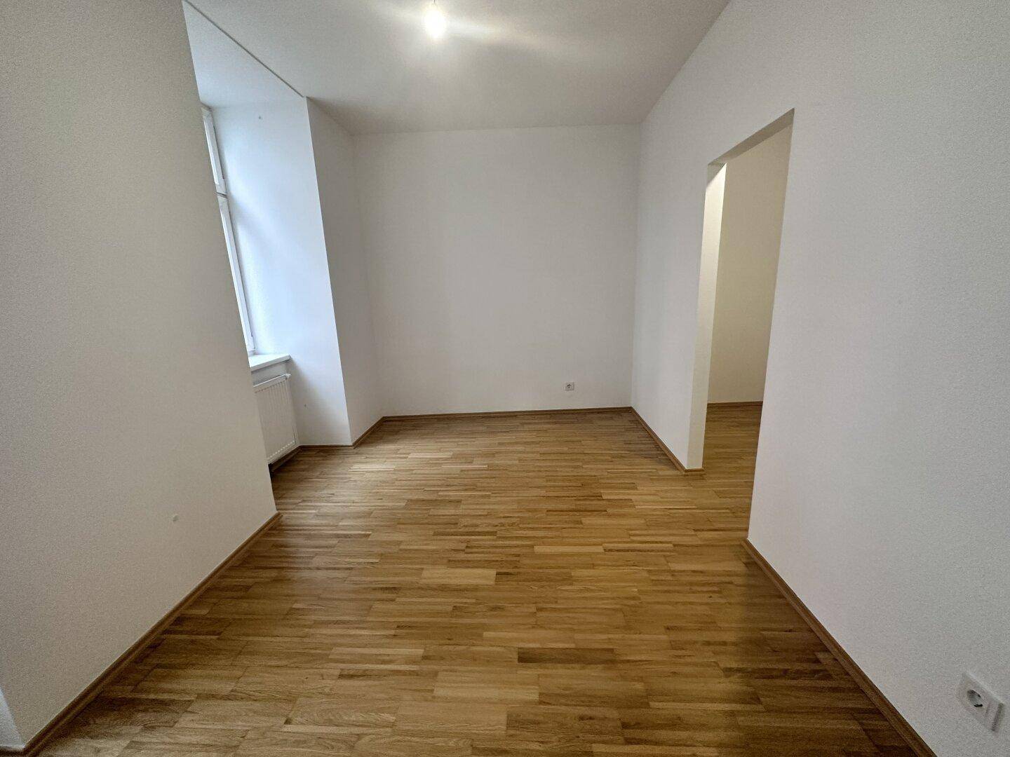 Geräumiges, leeres Zimmer mit hellem Parkettboden und einem Fenster, ideal für Wohnzwecke.