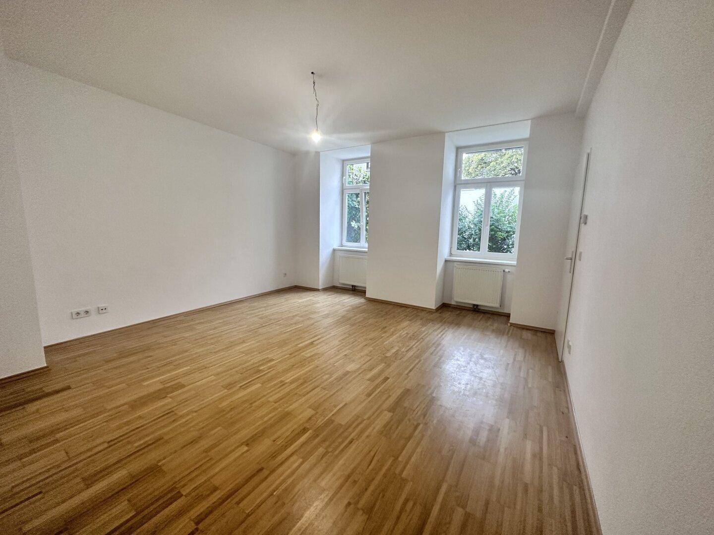 Helles, leeres Zimmer mit Parkettboden und zwei Fenstern, bereit zur individuellen Gestaltung.
