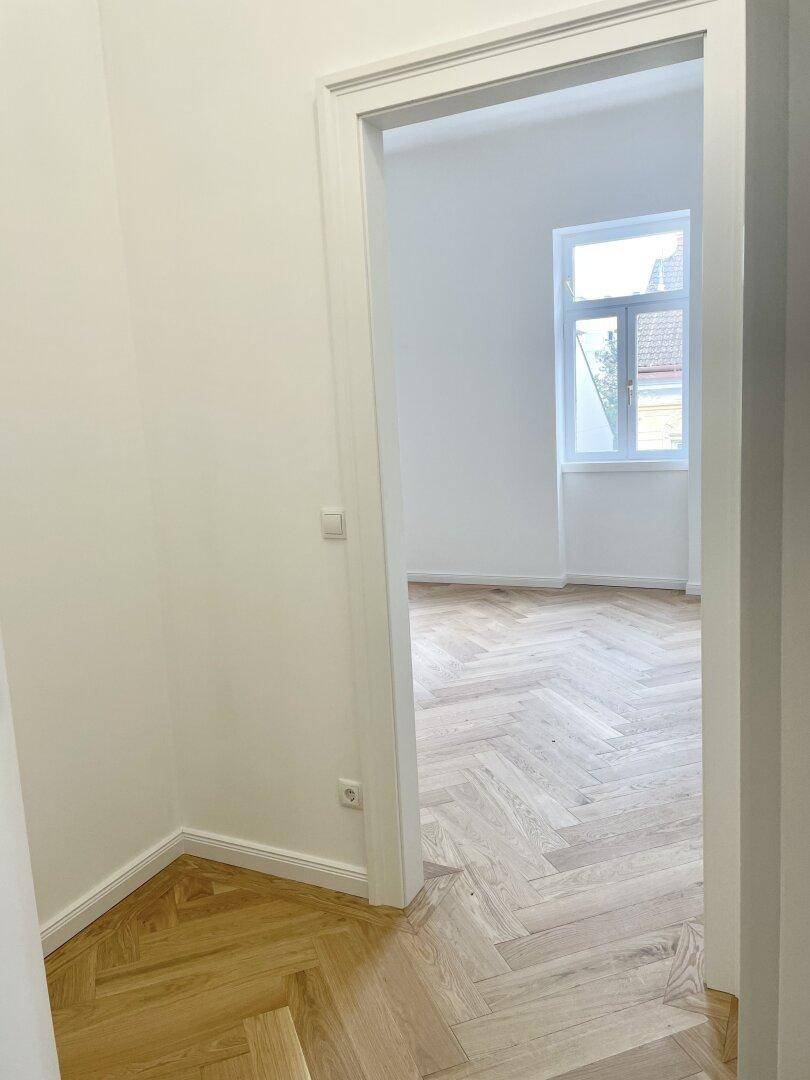 Blick durch eine Tür in einen hellen Raum mit Fischgrätparkett und einem Fenster, verbindet die Bereiche.