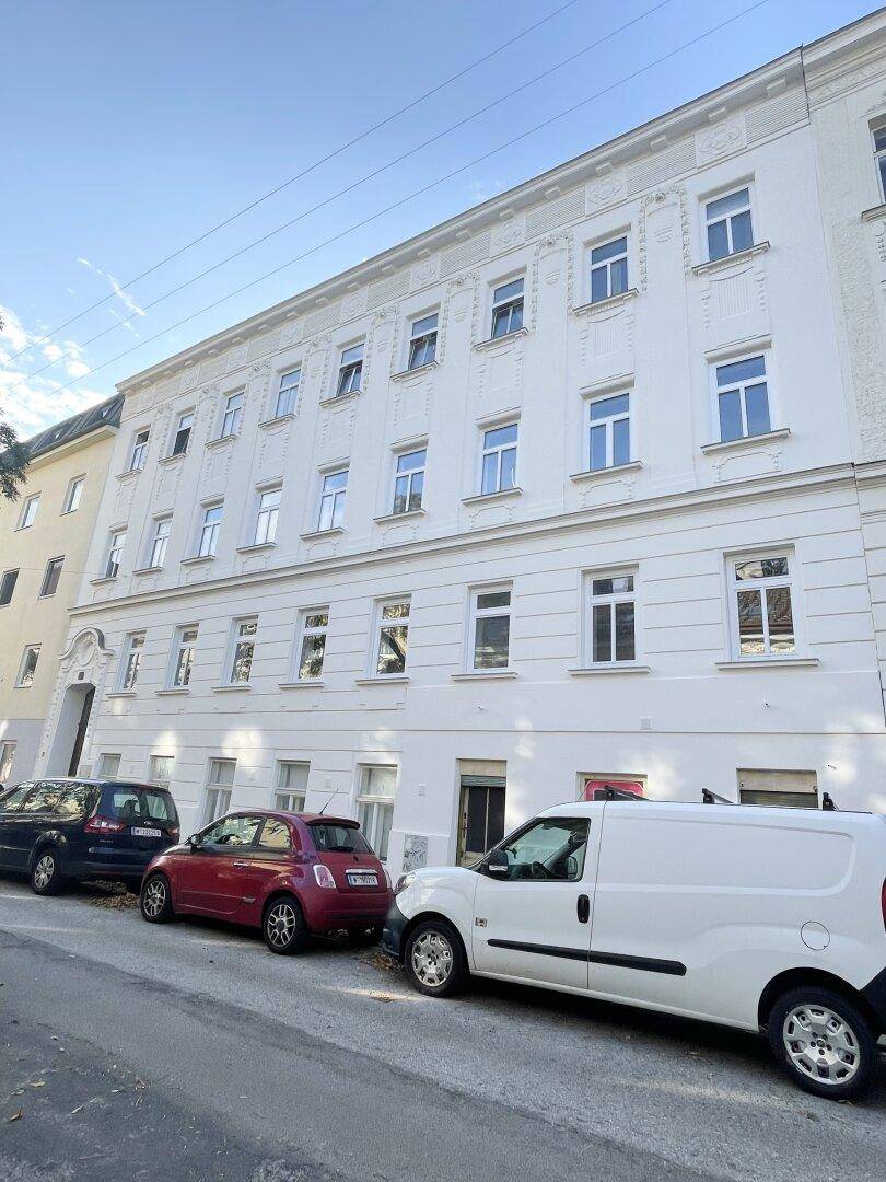 Ansicht der weißen Altbaufassade mit vielen Fenstern und parkenden Fahrzeugen auf der Straße.