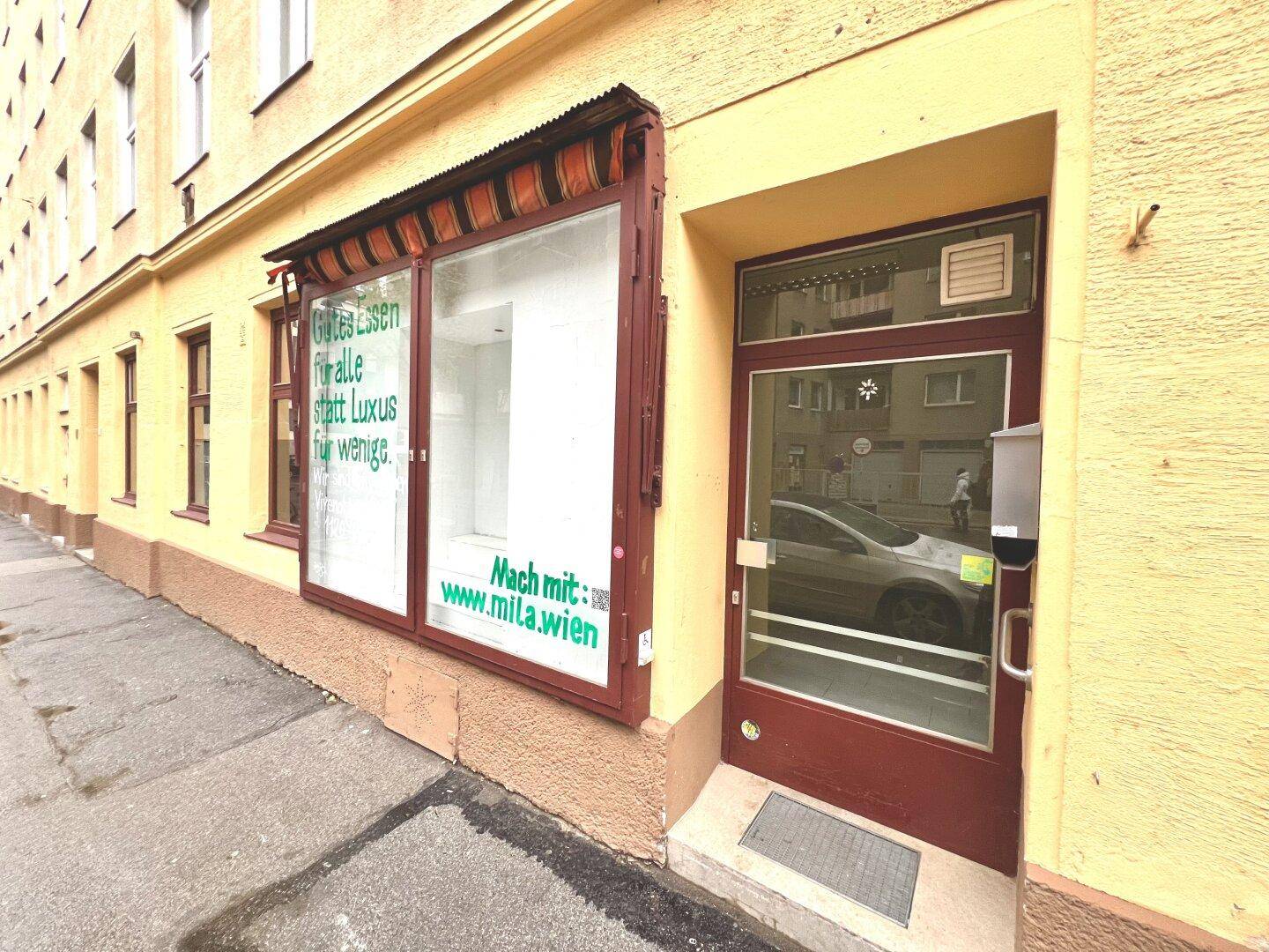 Eingangsbereich eines Geschäfts mit Schaufenstern und einer roten Tür an einer belebten Straße.